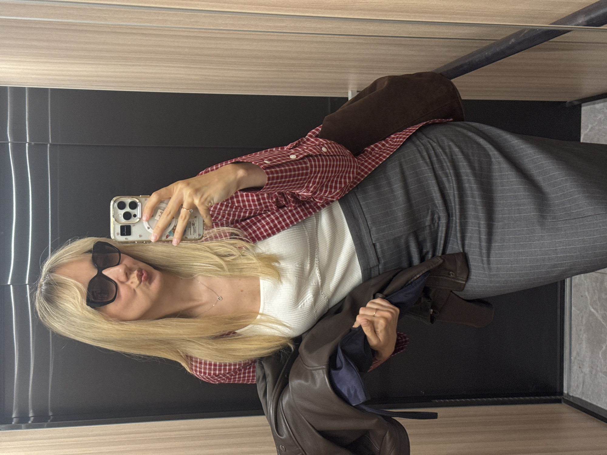 A woman with long, straight blonde hair and dark sunglasses is depicted taking a mirror selfie indoors. She holds a light-colored smartphone in her right hand, capturing her reflection, and poses with playfully pursed lips.
She is dressed in a red and white plaid button-down shirt worn over a white ribbed top. A delicate silver necklace with a small square pendant adorns her neck. A simple gold ring is visible on her left hand. Her lower attire consists of a dark gray garment with subtle pinstripes, possibly a skirt or wide-leg trousers. She holds a dark brown leather-like jacket with a blue lining draped over her left arm. A dark brown shoulder bag is slung over her right shoulder.
The setting features dark, reflective wall panels, likely a mirror, with lighter wooden panels above and a light-colored, possibly tiled floor. A dark metal bar or railing is visible in the upper part of the reflection. The environment suggests a modern indoor space, such as a hotel corridor or an elevator. The overall mood is casual and confident. While some text is visible on the phone case, it is illegible. The location context for the image is Riga, Latvia.