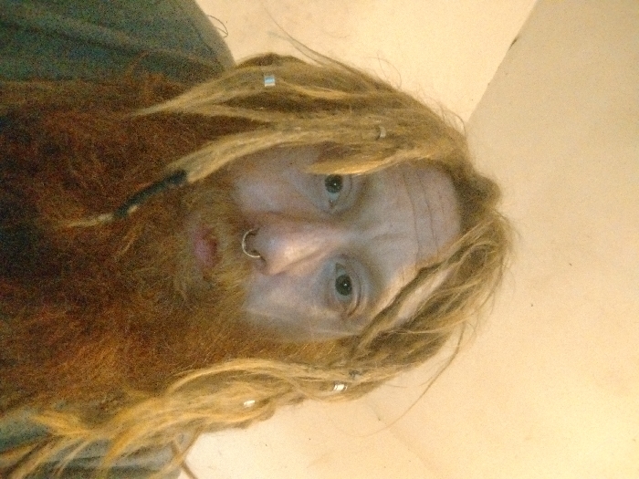 This close-up portrait depicts an individual, appearing male, with light eyes, a prominent reddish-brown beard, and long blonde dreadlocks, several of which are adorned with small silver-colored cylindrical beads. A silver septum ring is piercing the individual's nose. The subject is wearing a dark, likely olive-green, upper garment. The background is a plain, light-colored interior surface, possibly a wall or ceiling, showing minor textural imperfections. The exact location within Grand Rapids, United States, is not ascertainable from the visual information provided in the image.