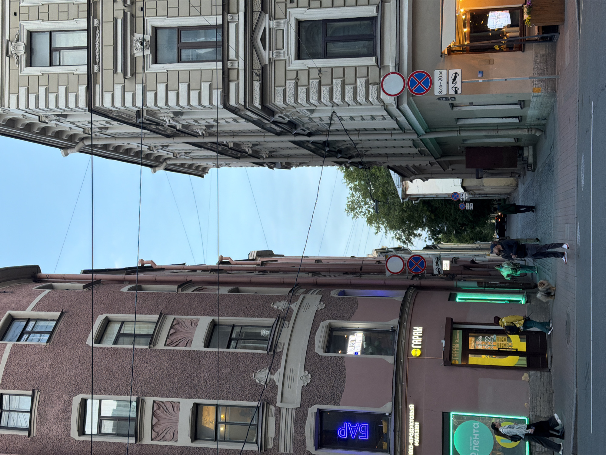 The image depicts a narrow street in Saint Petersburg, Russia, lined with multi-story buildings on either side. The building on the left is a salmon-pink color with ornate architectural details and multiple windows. Several signs are visible on its lower floors, including a neon blue sign that reads "БАР" (Bar) and a yellow sign for a store named "ГАРАЖ" (Garage) with operating hours. The building on the right is a pale beige color with a more classical architectural style, featuring decorative stone elements and numerous windows. Traffic signs are mounted on a pole on the right side of the street. Several people are present: two women walking on the sidewalk on the left, a man with a dog, and a few other figures further down the street. A small, fluffy dog is on a leash. The sky is overcast, suggesting late afternoon or early evening. Power lines are strung across the street, overhead.