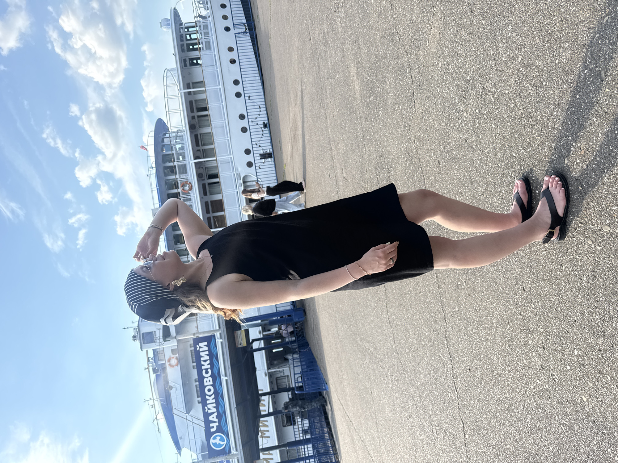 A full-body, side-profile shot captures a young woman standing on a paved surface in front of a large white passenger boat docked at a pier. The woman, who appears to be of East Asian descent, is wearing a black sleeveless dress, black flat sandals, a black and white striped bandana covering her hair, and large gold hoop earrings. She is looking up and to the right, with her right hand shielding her eyes from the sun. The boat is labeled with Cyrillic text that reads "ЧАЙКОВСКИЙ" (Chaykovskiy), indicating the location is the city of Chaykovsky, Russia. The boat has multiple decks and windows, and a red life preserver is visible on its side. The sky is blue with scattered white clouds. In the background, several other people are visible near the boat and on the pier. The ground is a rough, grey asphalt surface.