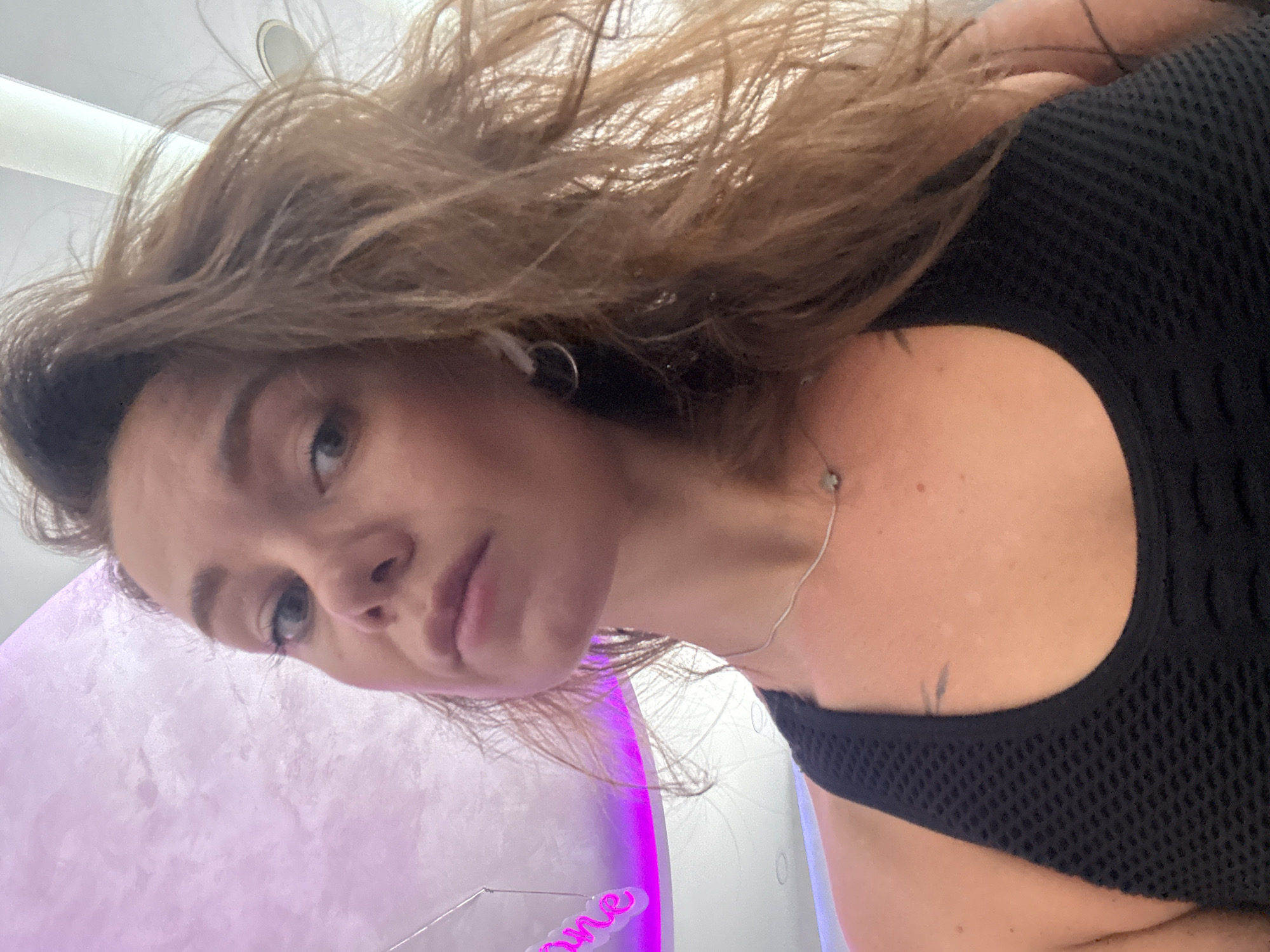 A woman with long, wavy brown hair and blue eyes looks towards the camera. She is wearing a black mesh tank top and a silver necklace with a small pendant. Her hair is windblown. Behind her, a pink neon sign with the word "one" is visible against a textured purple ceiling. The scene appears to be an indoor environment, possibly a gym or studio, with a bright light source above. The location is identified as Budva, Montenegro.