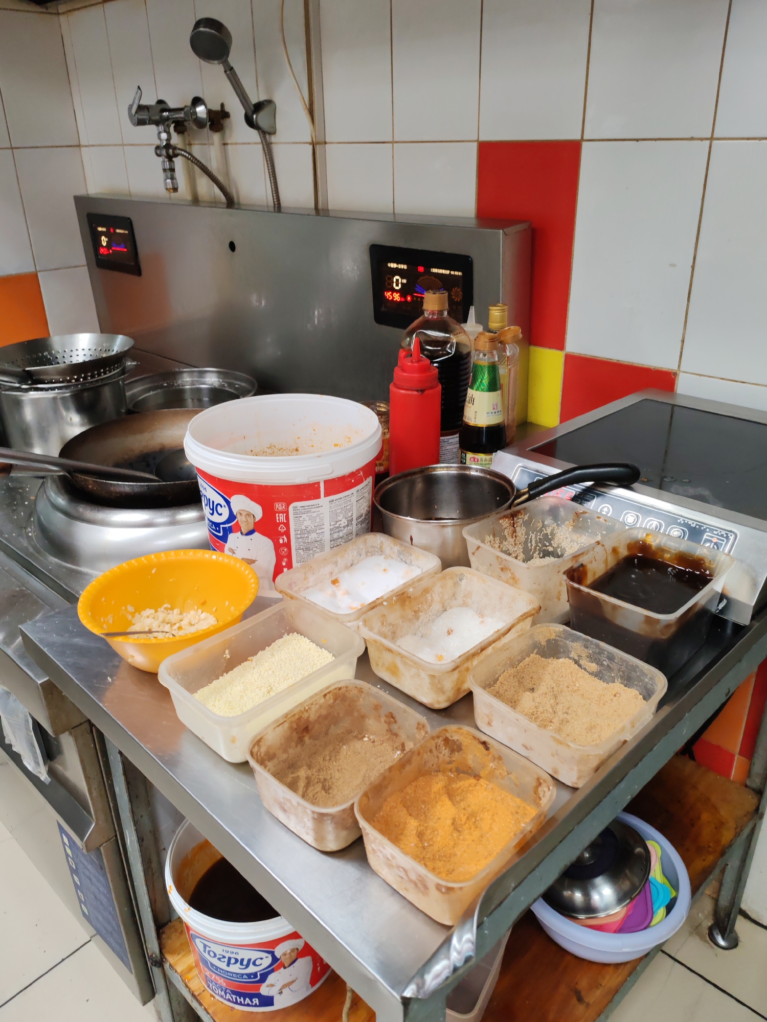 The image depicts a commercial kitchen setup, likely in Komsomolsk-on-Amur, Russia, judging by the Cyrillic text on the large white bucket. The scene is organized with various food ingredients and cooking equipment. In the foreground, a stainless steel counter holds several clear plastic containers filled with different powders and granular ingredients, possibly spices or breading. To the left, a yellow bowl contains small, light-colored food items, and a large white bucket with red and blue labeling, featuring an image of a chef and Cyrillic text, is also present. Behind these, a wok is positioned over a stovetop, suggesting active food preparation. In the background, a stainless steel unit with digital displays is mounted on the wall, along with a commercial faucet and spray nozzle. The tiled wall behind the equipment features alternating white and red squares. The overall impression is one of a functional, well-equipped kitchen preparing food.