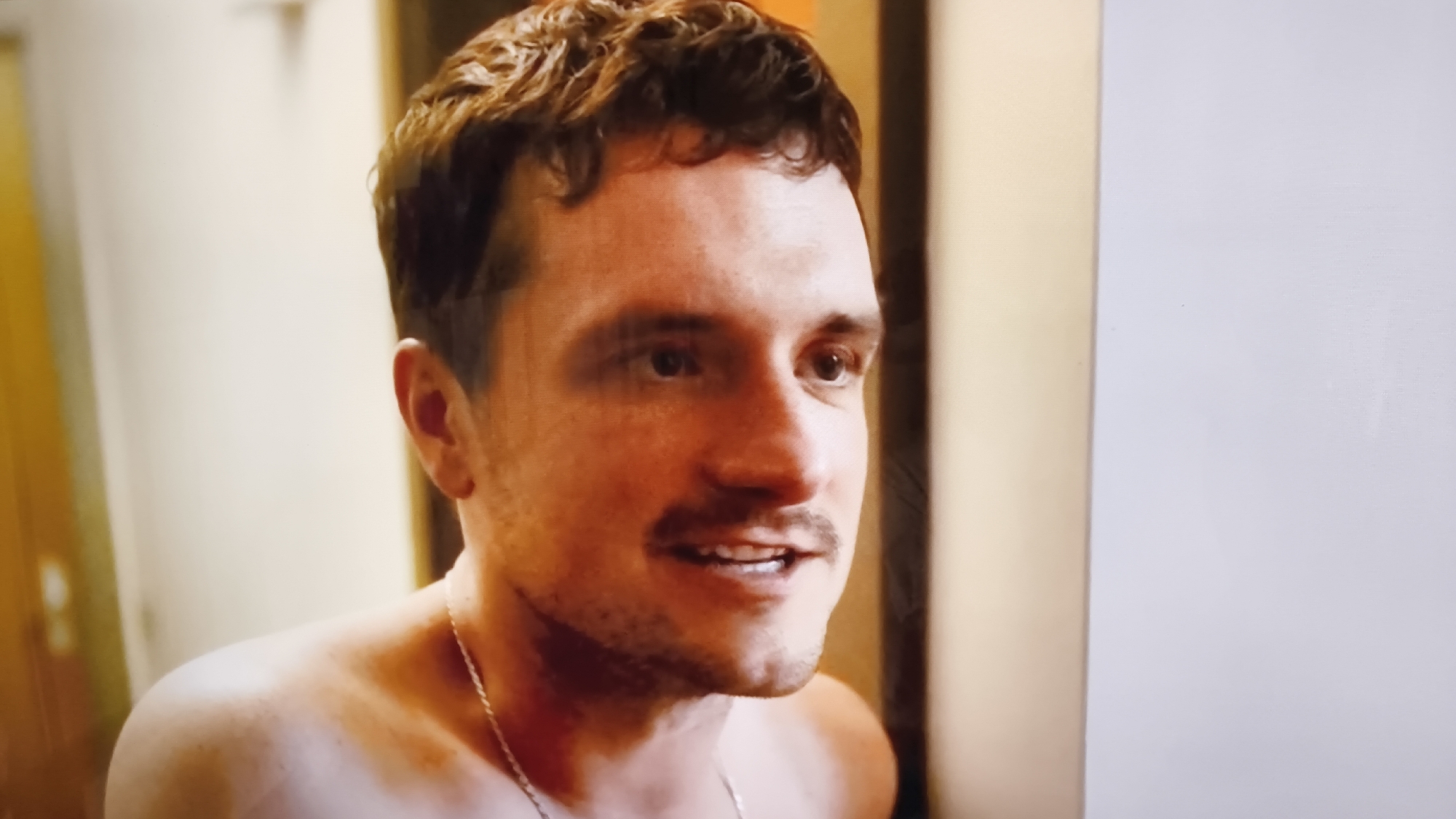 The image is a close-up of a man's upper body and face. He has short, dark, curly hair and a mustache. He is looking slightly off to the side with his mouth open as if speaking. He is wearing a thin silver chain around his neck. The background is out of focus, with a wall that appears to be a light color on the right side of the image and a darker, warmer color on the left. There is no indication of the city of Minsk, Belarus.
