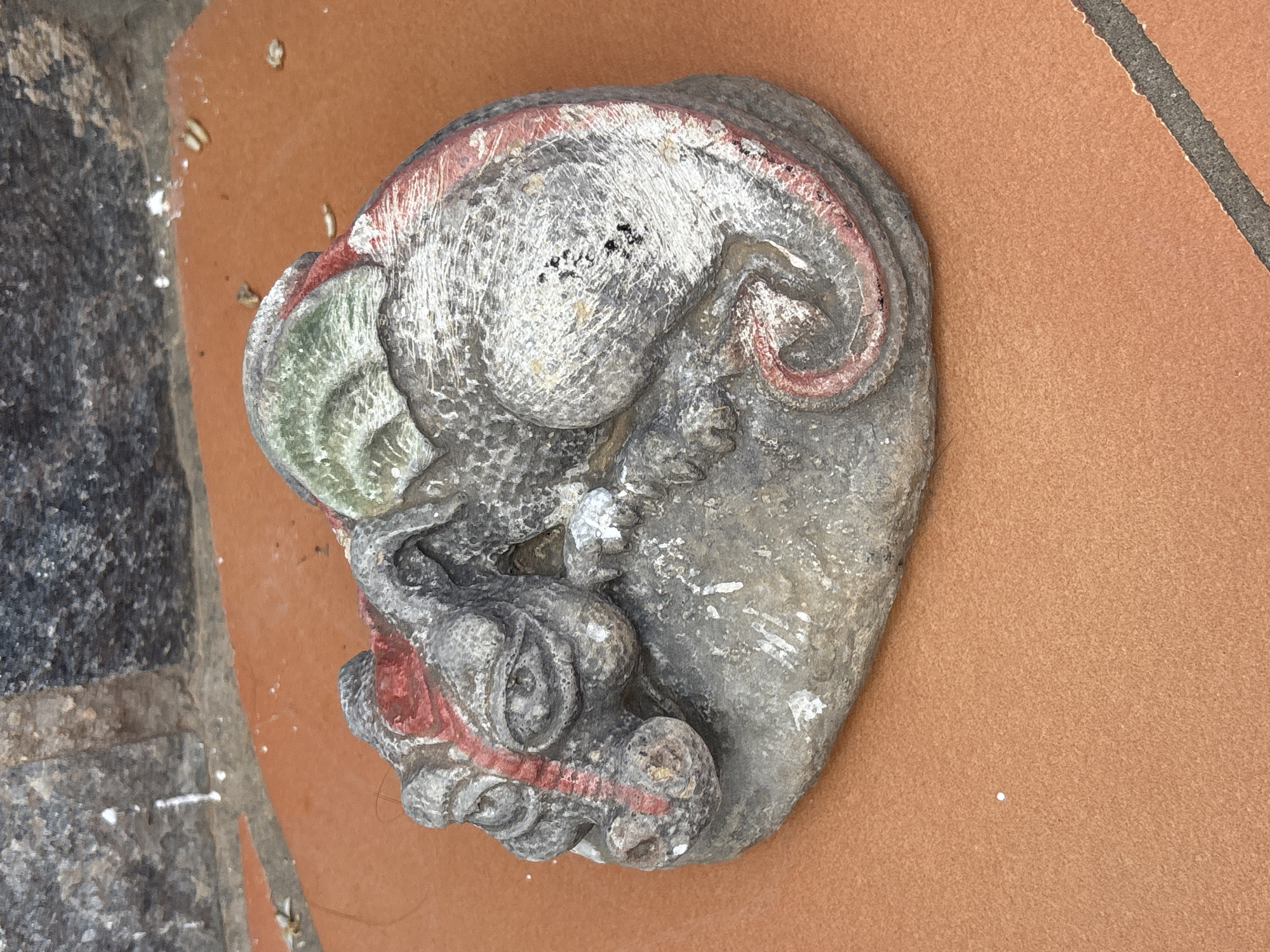 A small, weathered, grey stone sculpture of a coiled dragon with painted red and green accents rests on a textured, reddish-brown surface. The dragon's head faces left, with its eyes and snout visible. Red paint highlights its forehead and the edge of its tail. Green paint is visible on its wing. The sculpture is situated on what appears to be an outdoor tiled surface. In the background, a rough-hewn stone wall is visible, with some white debris scattered on the ground. The overall scene suggests an outdoor decorative element.
