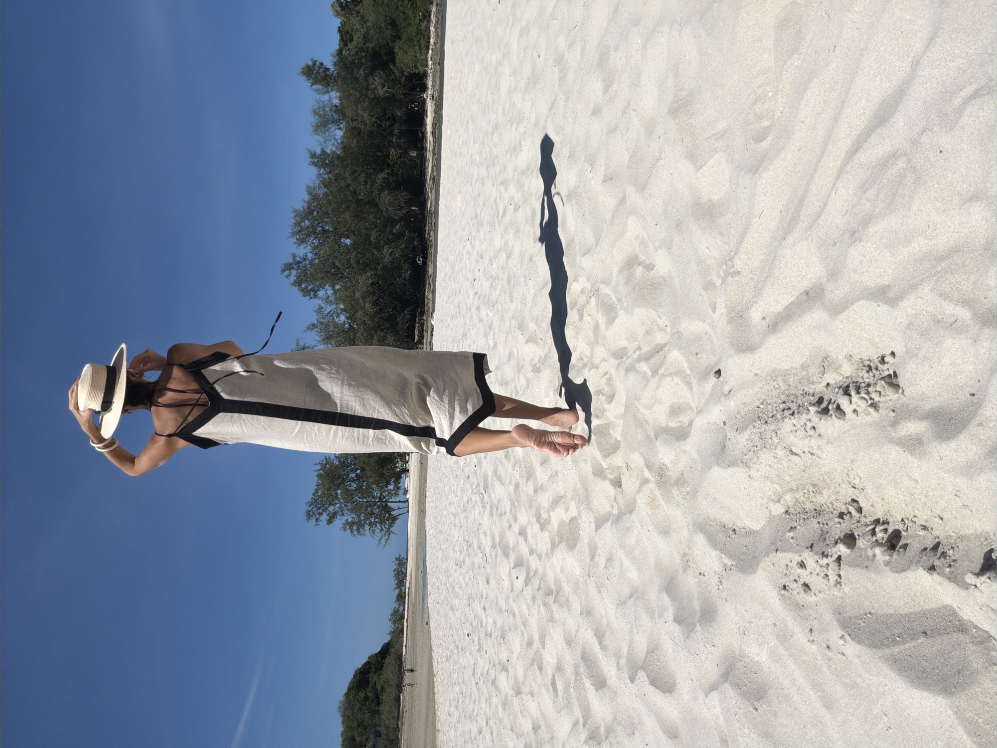 A woman wearing a straw hat and a long, light-colored dress with black trim stands on a sandy beach, facing away from the camera. She is adjusting her hat. Her bare feet are visible as she walks on the sand. The beach is wide and appears to be in a tropical location, with lush green trees and foliage in the background. The sky is a clear, bright blue. The scene suggests a leisurely day at the beach in Baan Plai Laem, Thailand.