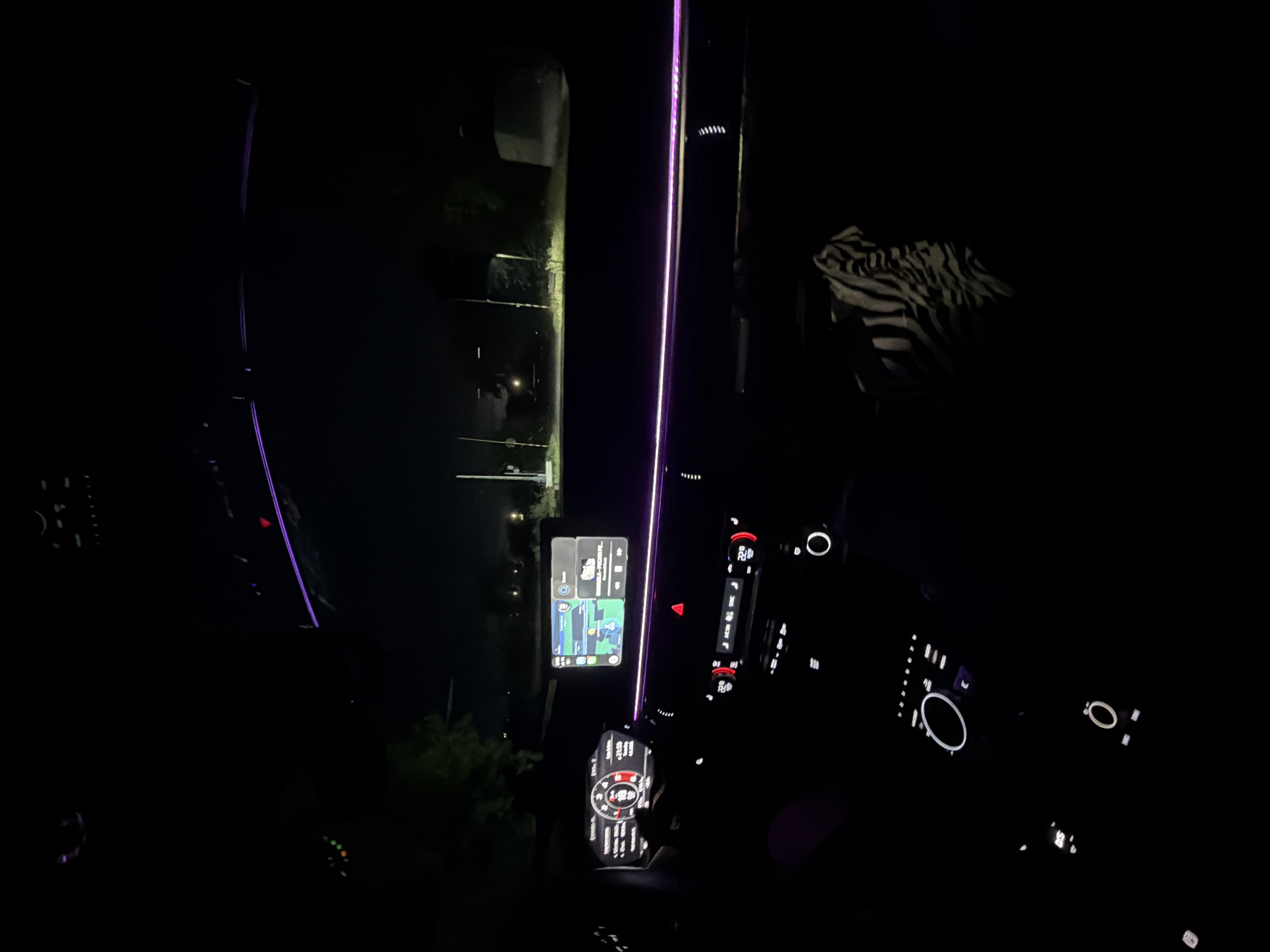 The image captures the interior of a car at night, looking out through the windshield onto a dark street in Pirgovo, Bulgaria. The primary subjects are the illuminated car dashboard and a section of the road ahead.
Inside the vehicle, prominent purple ambient LED lighting strips run along the top of the dashboard and down the driver's side door trim, casting a vibrant glow. The central infotainment screen, mounted vertically, displays what appears to be a navigation application, possibly Google Maps, showing a route and a dark map interface. Visible text on this screen includes a time "16:03," a weather icon with "22°C," and what looks like "Pirgovo" and other city names like "Burgas" and "Plovdiv" on the map. Below the infotainment screen is the digital instrument cluster, showing a speedometer with "120" prominently displayed, suggesting a speed of 120 km/h, and a digital clock reading "21:53." To the right of the central screen, climate control dials indicate "22.0°C" for both driver and passenger sides. Various other illuminated buttons and controls are visible on the dashboard and steering wheel.
Outside the car, the scene is very dark, with faint lights visible in the distance along what appears to be a rural road or path. Some indistinct shapes of trees or buildings can be vaguely made out against the night sky. On the far right side of the car interior, a crinkled item with a black and white zebra print pattern is visible, likely a blanket or fabric on the passenger seat.
The activity depicted is either driving or being parked in a car at night, with the navigation system active, suggesting travel. The time on the instrument cluster (21:53) confirms the nighttime setting.