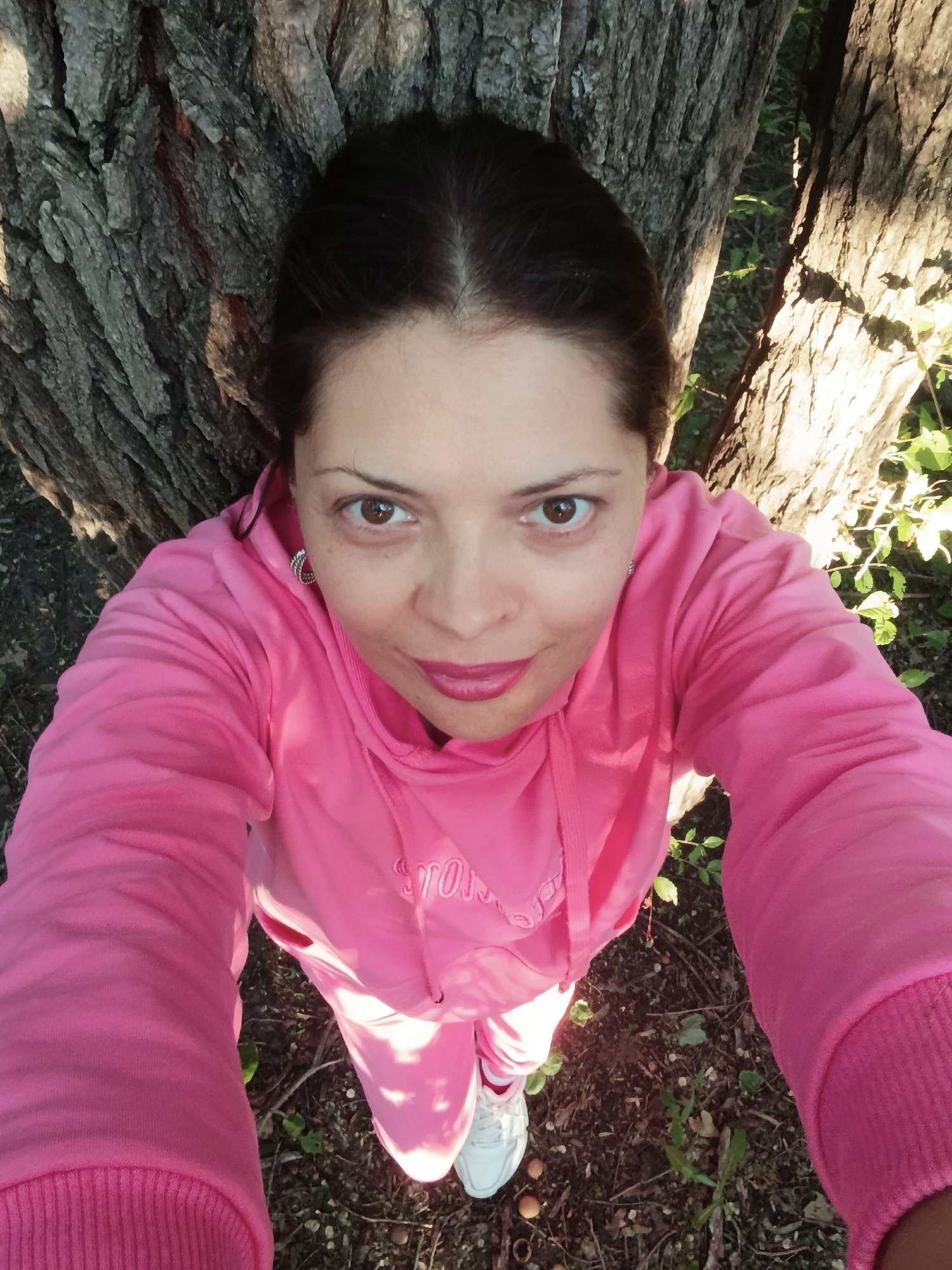 A high-angle, close-up selfie of a woman standing outdoors, leaning against a large tree trunk. The woman has dark hair, brown eyes, and is wearing a bright pink hooded sweatshirt and matching pants, along with white sneakers. She has on noticeable makeup, including bright pink lipstick, and is wearing a hoop earring. The background consists of the textured bark of a tree and some greenery on the ground. The lighting suggests it is daytime, with sunlight casting shadows and illuminating parts of the scene. There are no other people or distinct landmarks visible to indicate a specific location within Saratov, Russia.