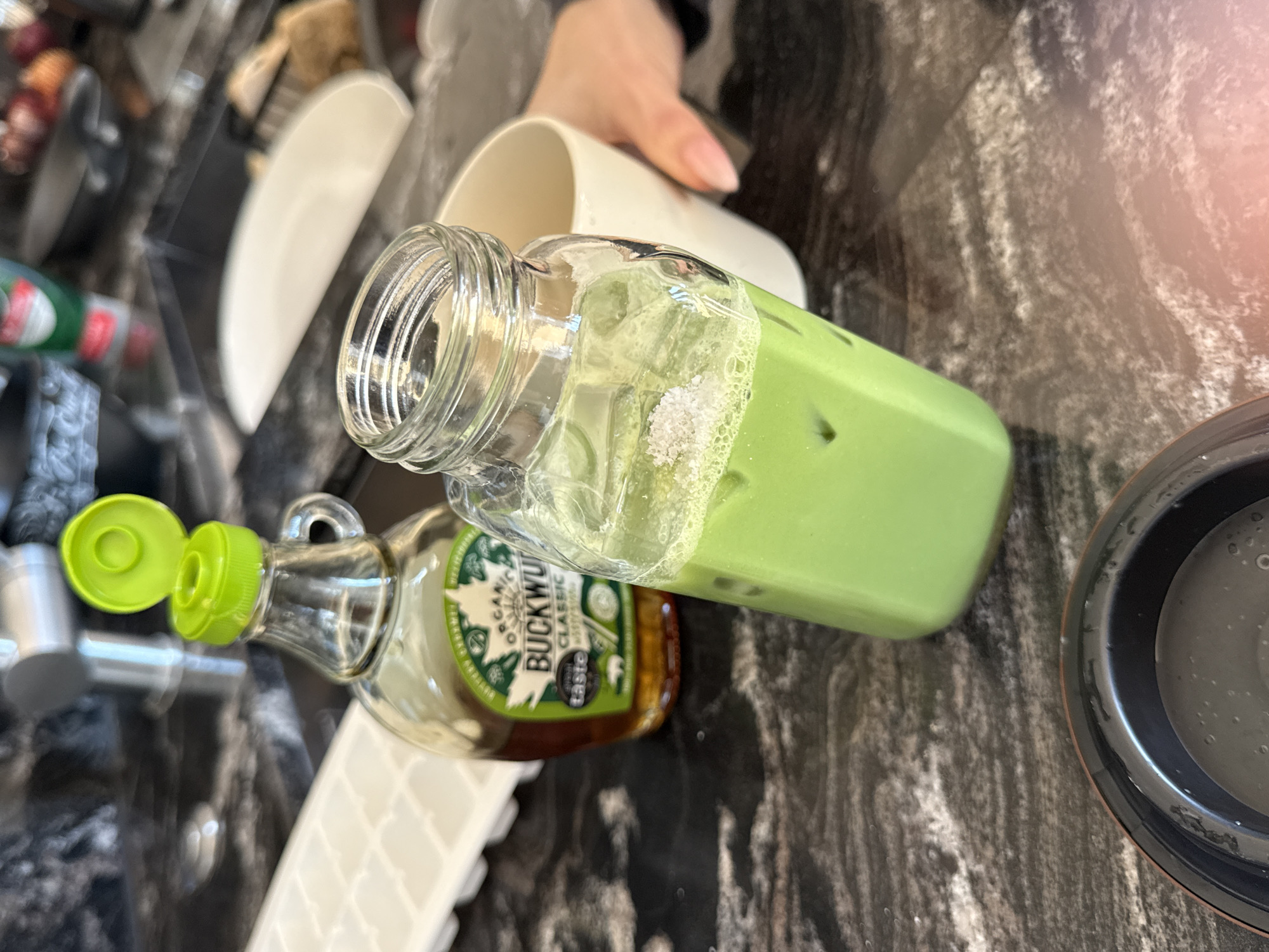 A close-up, elevated perspective photograph captures a domestic kitchen countertop scene. Dominating the center is a clear glass bottle filled with a vibrant light green liquid, consistent with an iced matcha latte, containing several ice cubes and a small cluster of white granular substance (likely sugar or salt) floating on its surface. To its left, a smaller clear glass bottle of amber-colored syrup, labeled "ORGANIC BUCKWHEAT CLASSIC," features a green label and an open lime-green flip-top cap. A white plastic ice cube tray is partially visible on the far left. In the blurred background, a person's right hand with light-colored painted fingernails holds a plain white ceramic mug. The scene unfolds on a dark, polished countertop, likely granite or marble, characterized by grey and white veining. A metallic faucet and other blurred kitchen items are discernible in the distant background. A dark, circular object with a bronze or copper rim is visible in the bottom foreground. There is no discernible information within the image to specify its location as the city of London, United Kingdom.
