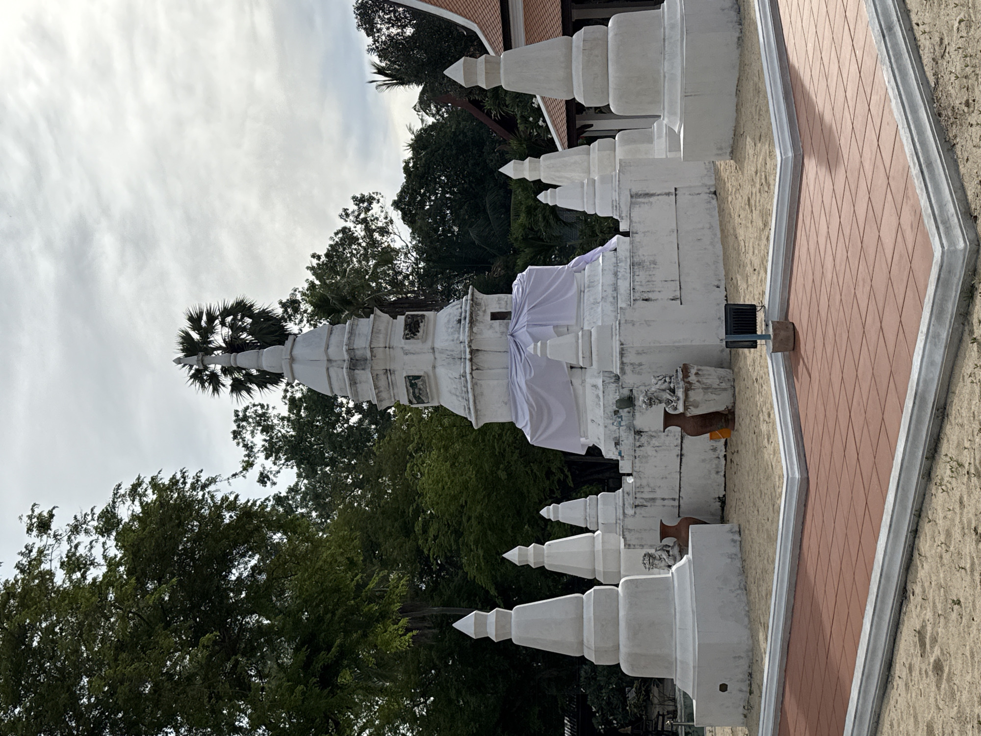 A white, tiered stupa is the central focus of the image, situated outdoors on a sandy area in Baan Tai, Thailand. The stupa is surrounded by several smaller, cone-shaped white structures. A white cloth is draped around the middle section of the main stupa. Two large, dark terracotta pots are visible near the base of the stupa, and a small dark square object, possibly a light fixture, is placed on a concrete base in the foreground. The background is filled with lush green trees and a cloudy sky. To the right, a portion of a building with an orange roof is visible. The ground in the foreground is a paved area with reddish-brown square tiles, bordered by a white concrete edge. The overall scene suggests a tranquil, possibly religious, outdoor setting.