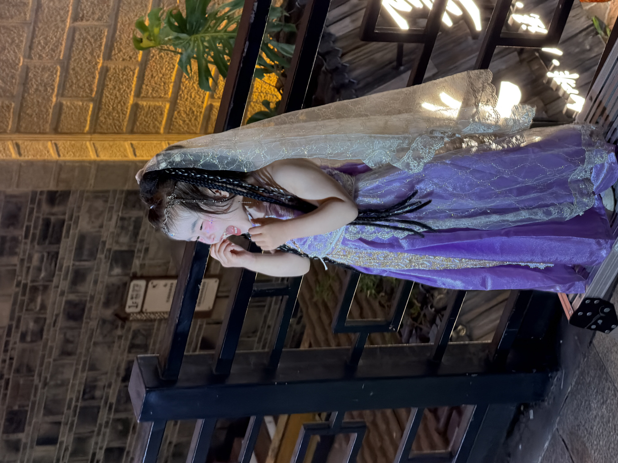 A young girl, wearing a purple dress and a veil, stands on a platform next to a railing in Yuzhong District, China. She has her hands near her mouth and her eyes are closed, suggesting she is laughing or singing. The background shows a brick wall and some greenery. The lighting suggests it is evening or night.