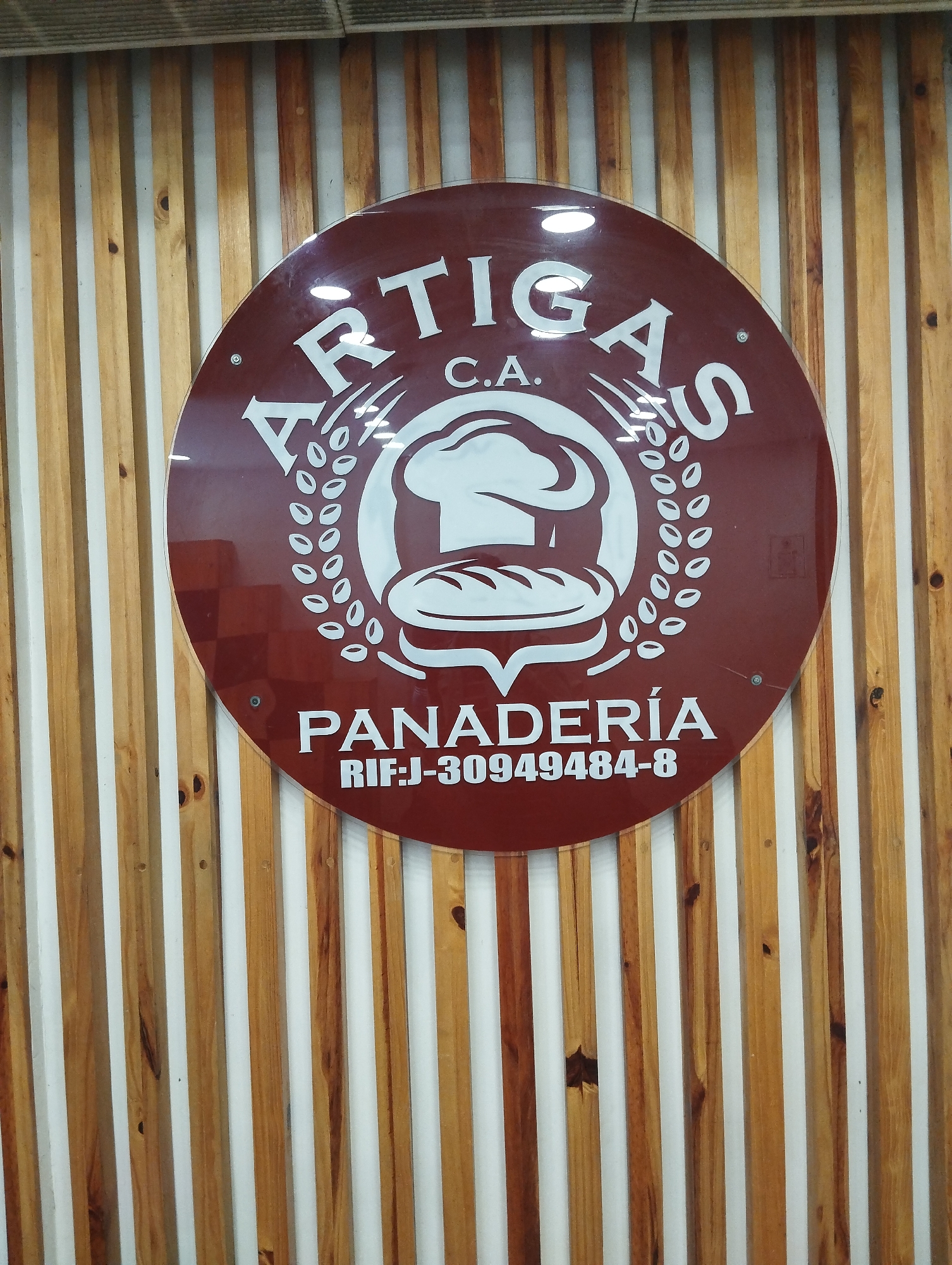A medium shot of a sign for a bakery called "Artigas, C.A." which is hung on a wall with vertical wooden slats. The sign is circular and maroon. The sign contains a logo consisting of a chef's hat and bread surrounded by wheat. The text on the sign says "Artigas C.A." at the top and "Panaderia RIF:J-30949484-8" at the bottom. The location is Porlamar, Venezuela. There are no people visible.