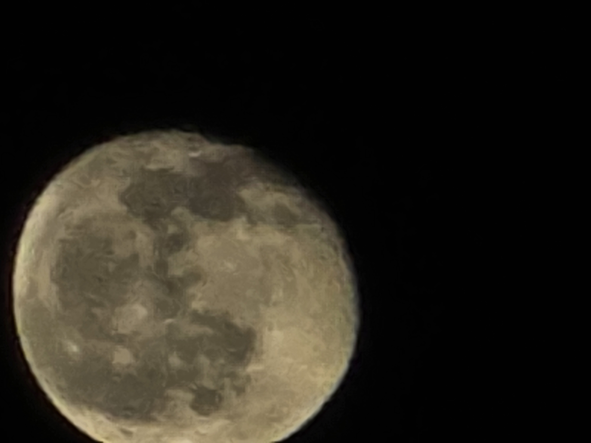 The image is a close-up, slightly blurry photograph of the full moon against a black, starless sky. The moon is illuminated and displays visible craters and surface features, appearing in shades of yellow and grey. The composition is such that the moon occupies the left half of the frame, with the right half being entirely black. The image is taken from an outdoor environment, likely at night, and there is no information available to specify its location in the city of Yambol, Bulgaria.