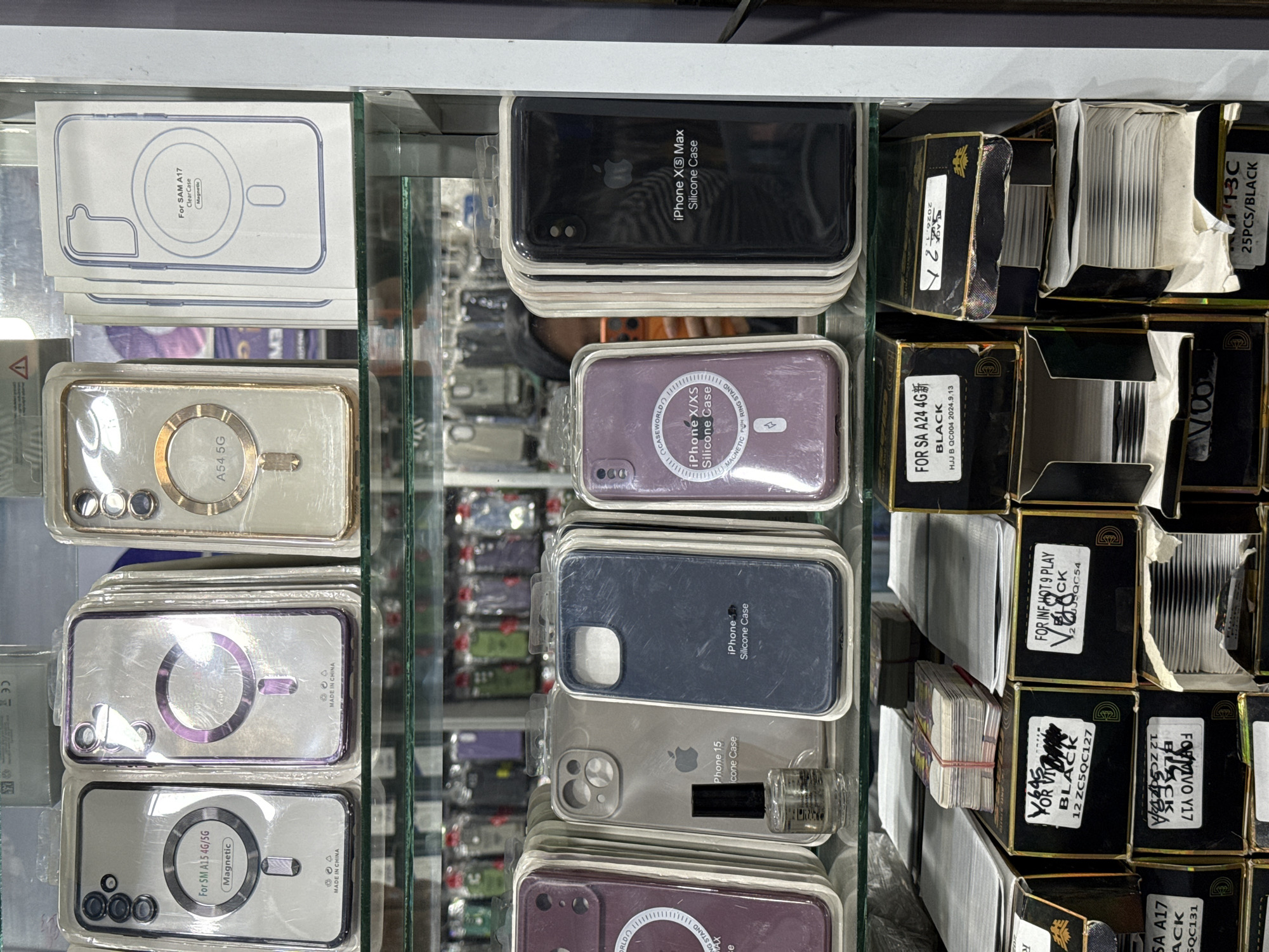 The image shows a retail display of phone cases in Bireuenmeunasahreuleut, Indonesia. The display is organized on multiple shelves within a glass-fronted cabinet. The top shelves feature various phone cases in their packaging, many designed for specific phone models such as "For SM A15 4G/5G," "A54 5G," and "For SAM A17 Clear Case." Some cases are transparent with magnetic rings, while others are colored silicone.
The middle shelves continue the display of phone cases, with some for "iPhone 15 Silicone Case" and "iPhone X/XS Silicone Case." A small glass bottle, possibly containing a cleaning solution or accessory, is placed next to one of the iPhone cases. The bottom shelves showcase stacks of boxes, some labeled with product names and quantities, such as "FOR SA A24 4G" and "25PCS/BLACK." The boxes appear to contain more phone cases or related accessories.
The overall environment suggests a mobile phone accessory shop. The lighting is adequate, highlighting the products on display. No people are prominently featured in the image, though a partial reflection of a person is visible in the glass cabinet on the middle shelf. The arrangement of items is neat and organized, indicating a typical retail setting.