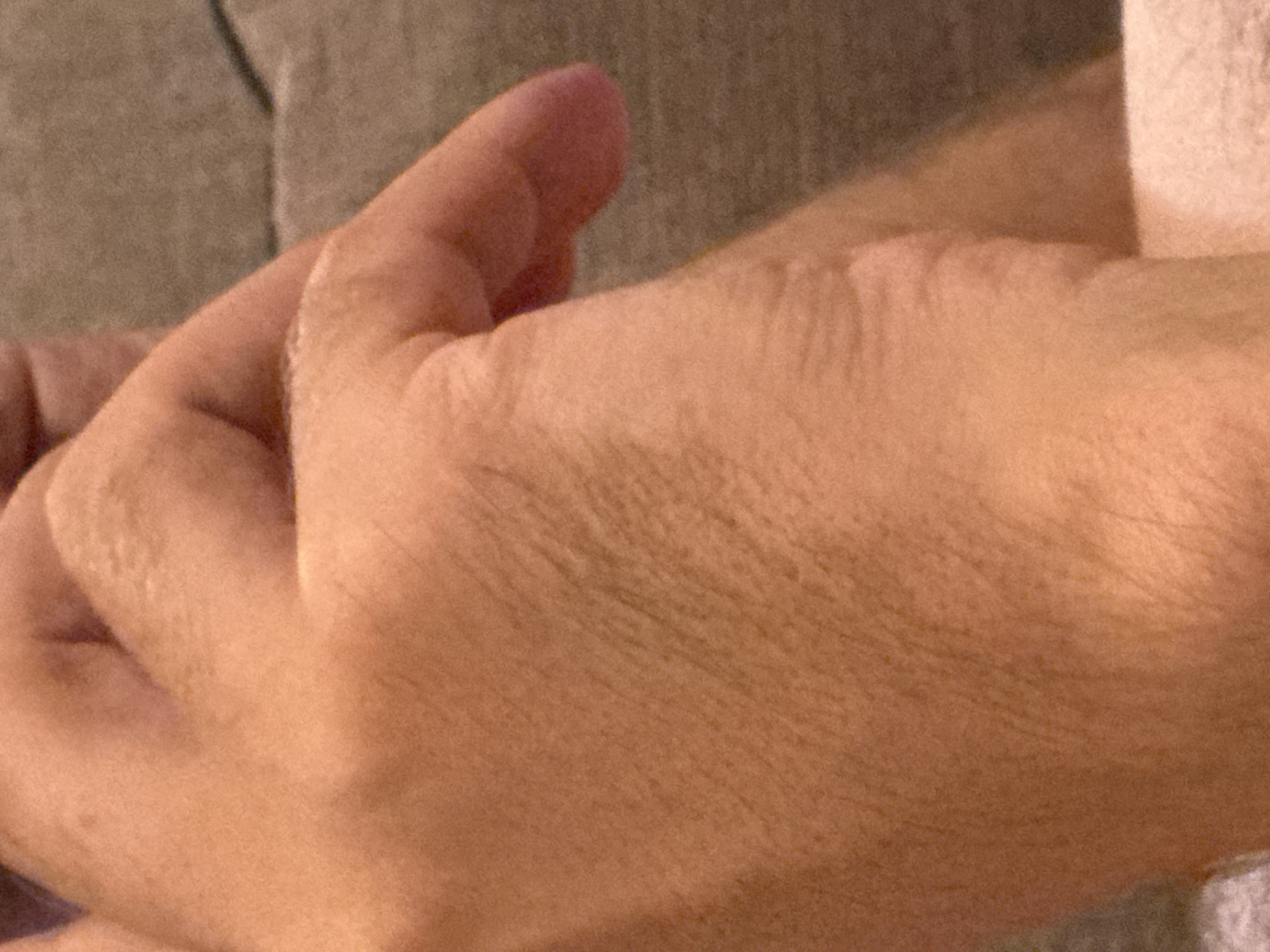 The image shows a close-up view of a person's hands, with a focus on the skin texture and the way light plays on the contours of the fingers and palm. The skin appears to be of a warm, tanned tone, with visible fine lines and pores. The lighting suggests a soft, ambient source, casting gentle shadows that emphasize the dimensionality of the hands. The background is blurred and appears to be a neutral, textured surface, possibly fabric or upholstery, in muted tones of brown and gray. No specific location in Budva, Montenegro, can be identified from this image. The overall impression is one of a still, intimate moment captured through a close examination of human anatomy.