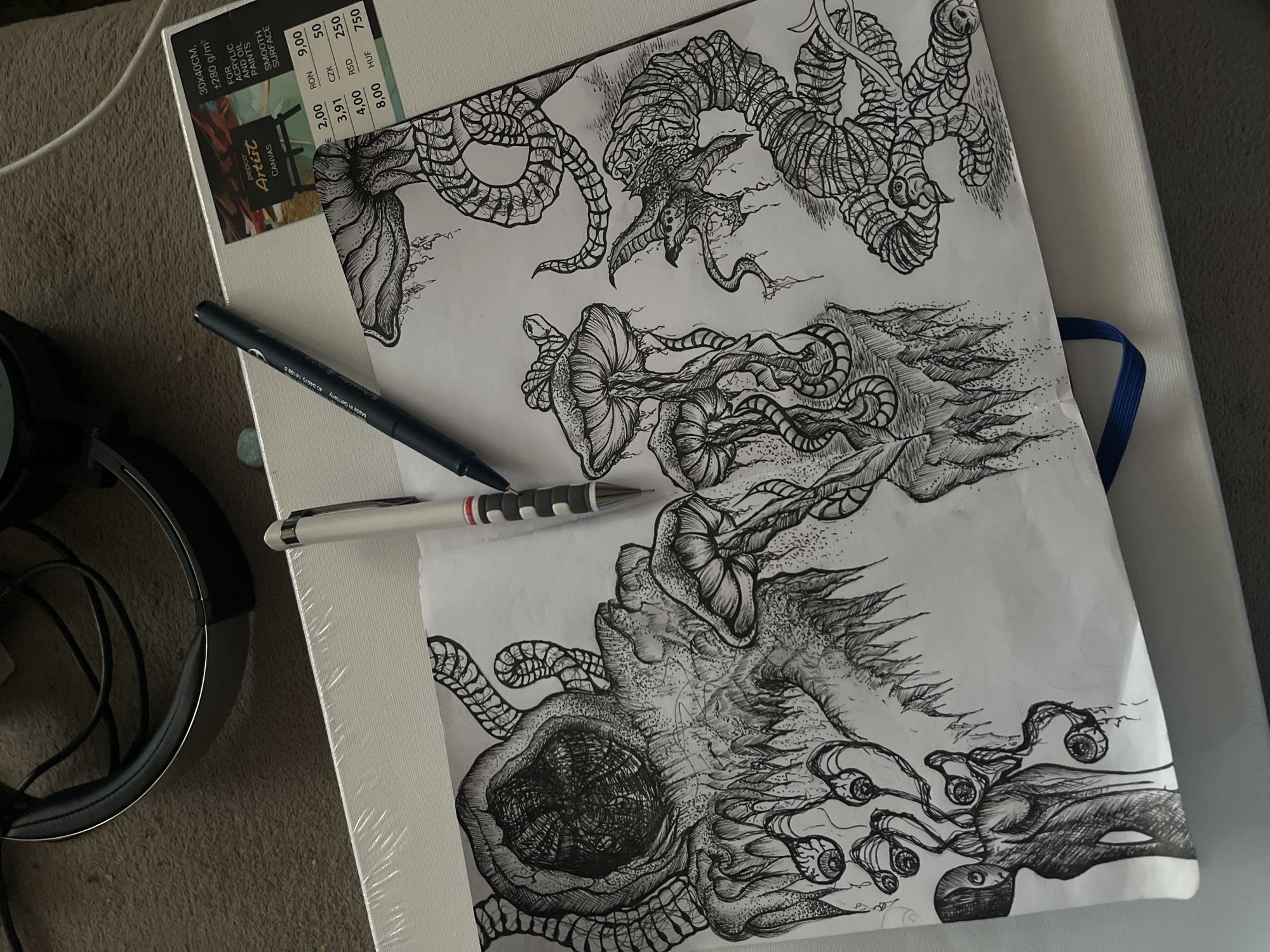 The image shows a white artist's canvas on which a black and white drawing is displayed. The drawing features several monstrous figures with tentacles and various strange appendages, including a creature with many eyes. Resting on the canvas and partially obscuring the artwork are two pens, one a blue rollerball and the other a white and black mechanical pencil. In the background, to the left, are a pair of black over-ear headphones. The canvas sits atop what appears to be a light grey surface. A portion of the canvas packaging is visible, indicating it is a "pepco Artist Canvas" with dimensions 30x40cm. There is no indication of this image being in Sofia, Bulgaria.