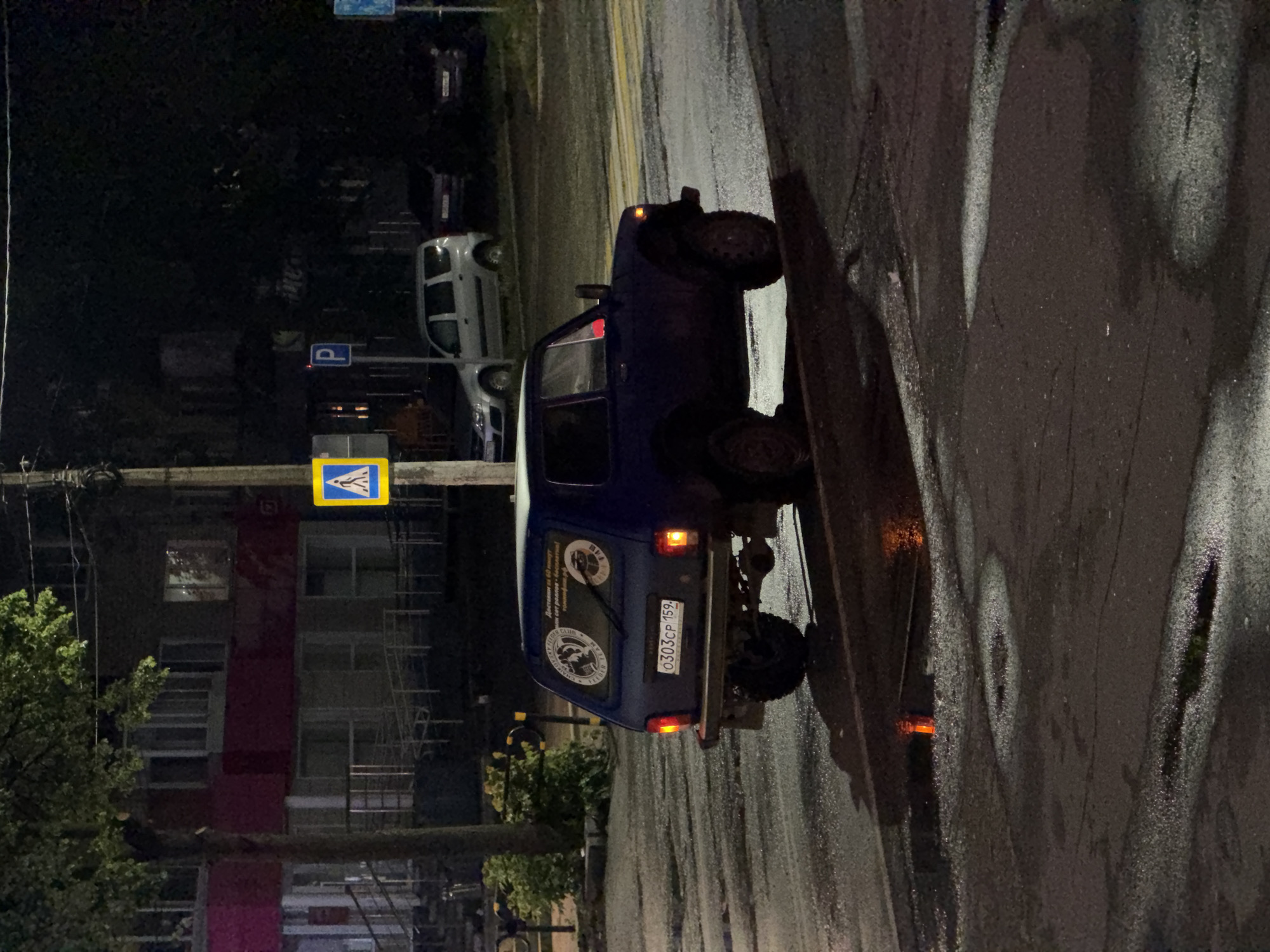 The image captures a night scene on a wet street, likely after rain, in an urban or suburban area. The main subject is a dark blue Lada Niva SUV, parked on the right side of the road. Its rear lights are illuminated, and it features large, possibly off-road, tires.
The license plate "0303CP 159" is clearly visible on the Lada Niva. On the car's rear window, a rectangular sticker displays Russian text, including "Доставка за 60 минут" (Delivery in 60 minutes) and "сет роллов бесплатно!" (free roll set!). Two round, black and white stickers are also present; one features what appears to be a bear head, and the other has text that includes "ER TEAM." The OCR also picked up "CHAIKOVSKY," indicating the city context.
In the background, on the left side of the street, other cars are parked, including a white sedan. Several street signs are visible, including a blue pedestrian crossing sign (featuring walking figures) and a blue parking sign with a white 'P'. A multi-story building with a prominent red vertical stripe stands in the distance on the left. Trees and artificial street lighting illuminate the damp environment, casting reflections on the wet road. The overall impression is a quiet street scene at night.