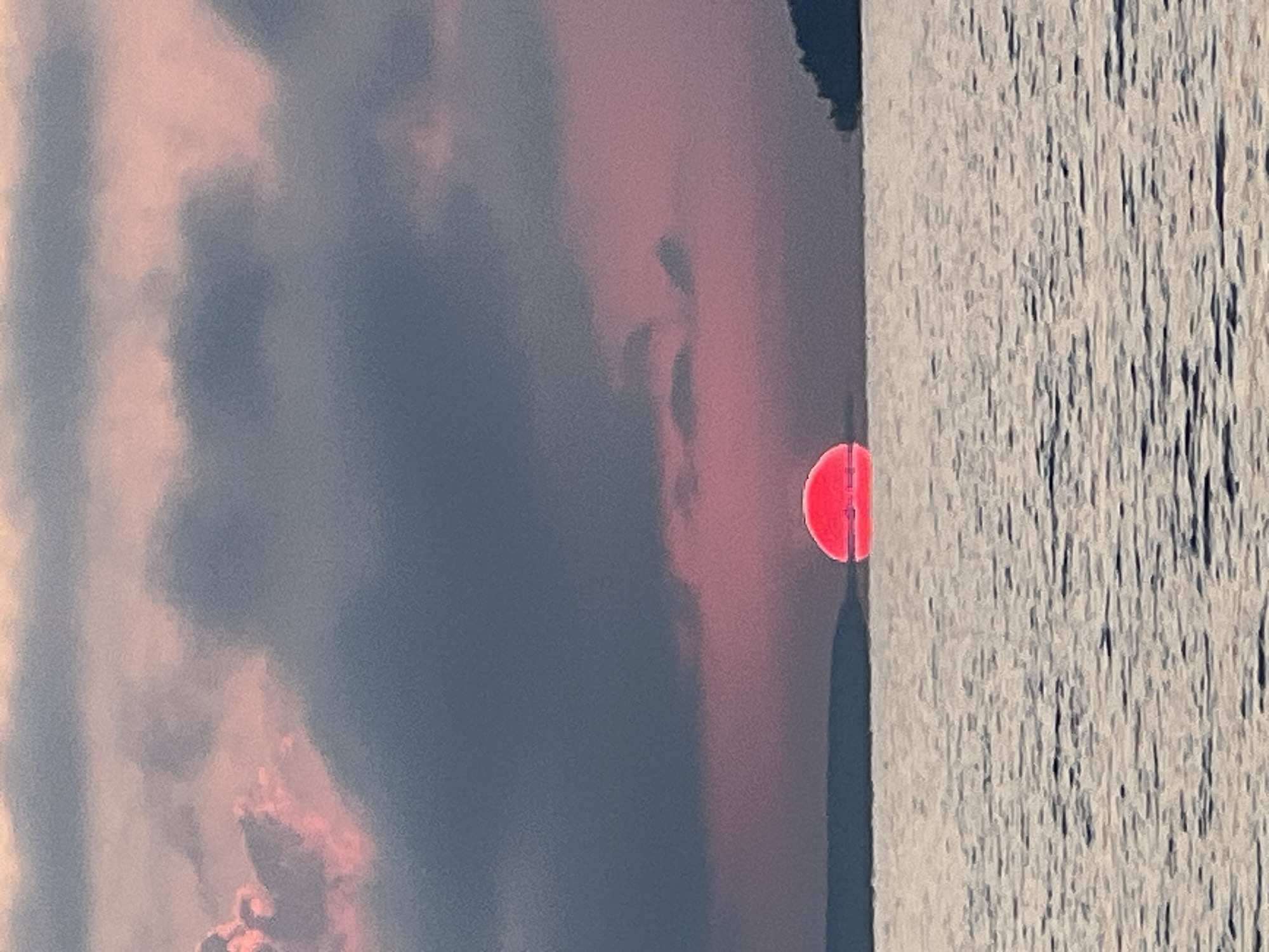 The image shows a sunset over a body of water, likely a lake or sea, with landmasses in the distance. The sun, a vibrant pink orb, is partially submerged at the horizon, casting a warm glow. The sky is filled with a mix of dark, moody clouds and softer, pink-tinted clouds, suggesting a dramatic atmospheric event. The water is calm, with ripples reflecting the diffused light. The overall scene evokes a sense of tranquility and natural beauty. This image does not contain any information that specifies its location in Novopichugovo, Russia, or any other city.