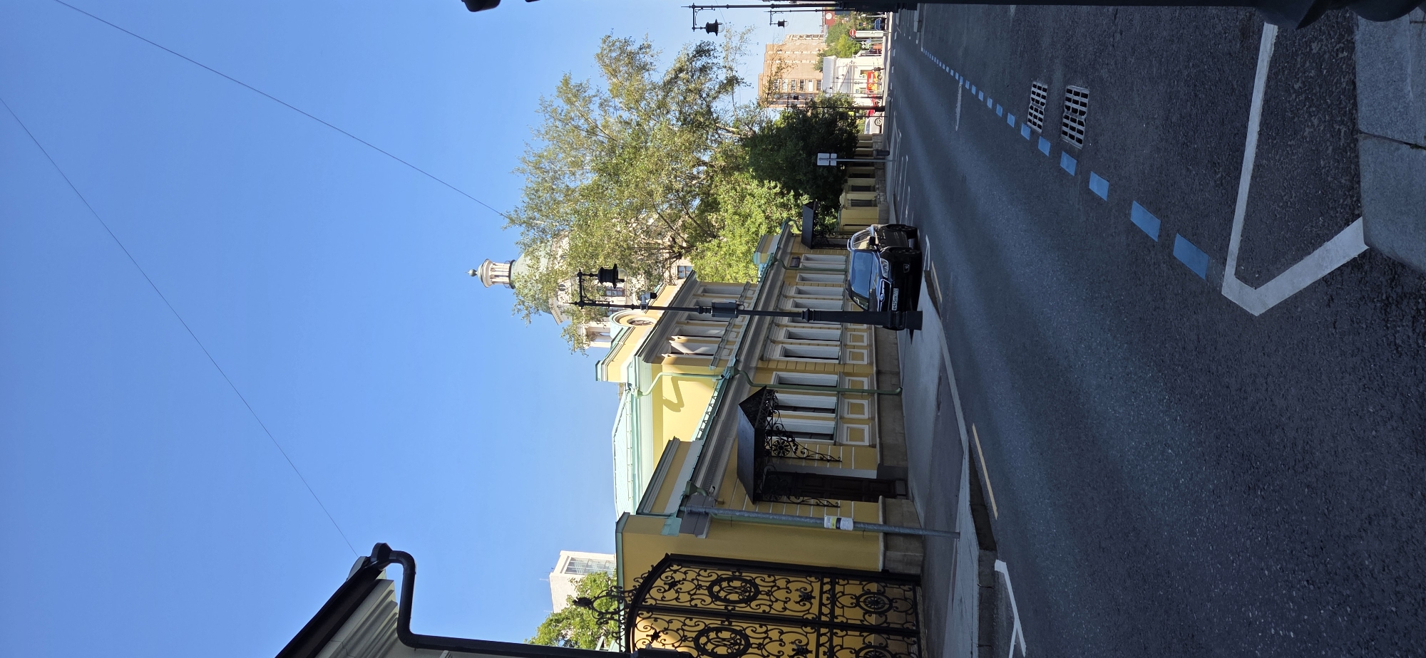 A black Subaru Forester is parked on the side of a street in Moscow, Russia. The car is facing away from the viewer, and its license plate is visible. The street is paved with asphalt and has white and blue markings. On the left side of the street, there is a yellow building with a large ornate black gate. Behind the building, a large tree partially obscures a green-domed building with a white tower. The sky is clear and blue.