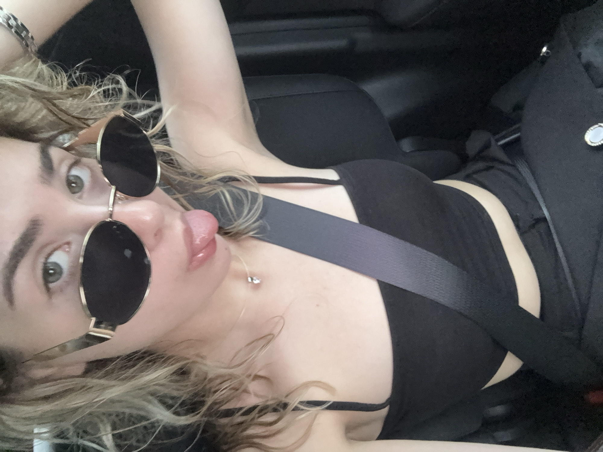 The image displays a head-and-torso selfie of a young female, reportedly taken in Sofia, Bulgaria, from the passenger seat of a vehicle. She has light-colored wavy hair and light-colored eyes, partially obscured by oval-shaped sunglasses with dark lenses and thin gold-toned frames. Her left eye is visible above the sunglasses. She is looking directly at the camera with her tongue extended. She wears a black spaghetti-strap crop top, and a dark grey seatbelt is fastened diagonally across her chest and lap. Accessories include a delicate necklace with a small, clear faceted pendant, and a chunky silver-toned link bracelet on her raised left wrist. A ring with a light-colored oval stone, possibly blue or white, set in metal, rests on a dark surface near her lap. The vehicle's interior appears dark with black or dark grey upholstery visible on the seat and headrest to her right. The scene is illuminated by natural light, indicating daytime.