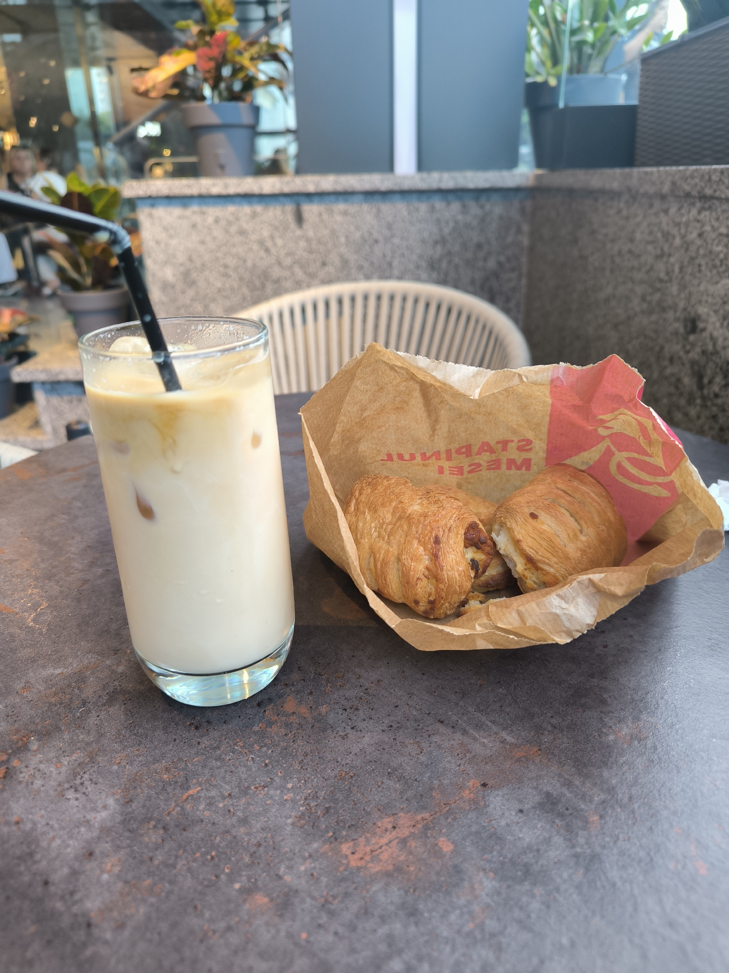 An iced beverage in a tall glass with a black straw sits on a dark, textured surface, next to a brown paper bag containing two pastries. The bag has red printing on the inside. In the background, out of focus, are plants in pots, a chair, and a wall. The overall scene suggests an outdoor seating area of a cafe. There are no identifiable people in the image. The location is not specified in the image.