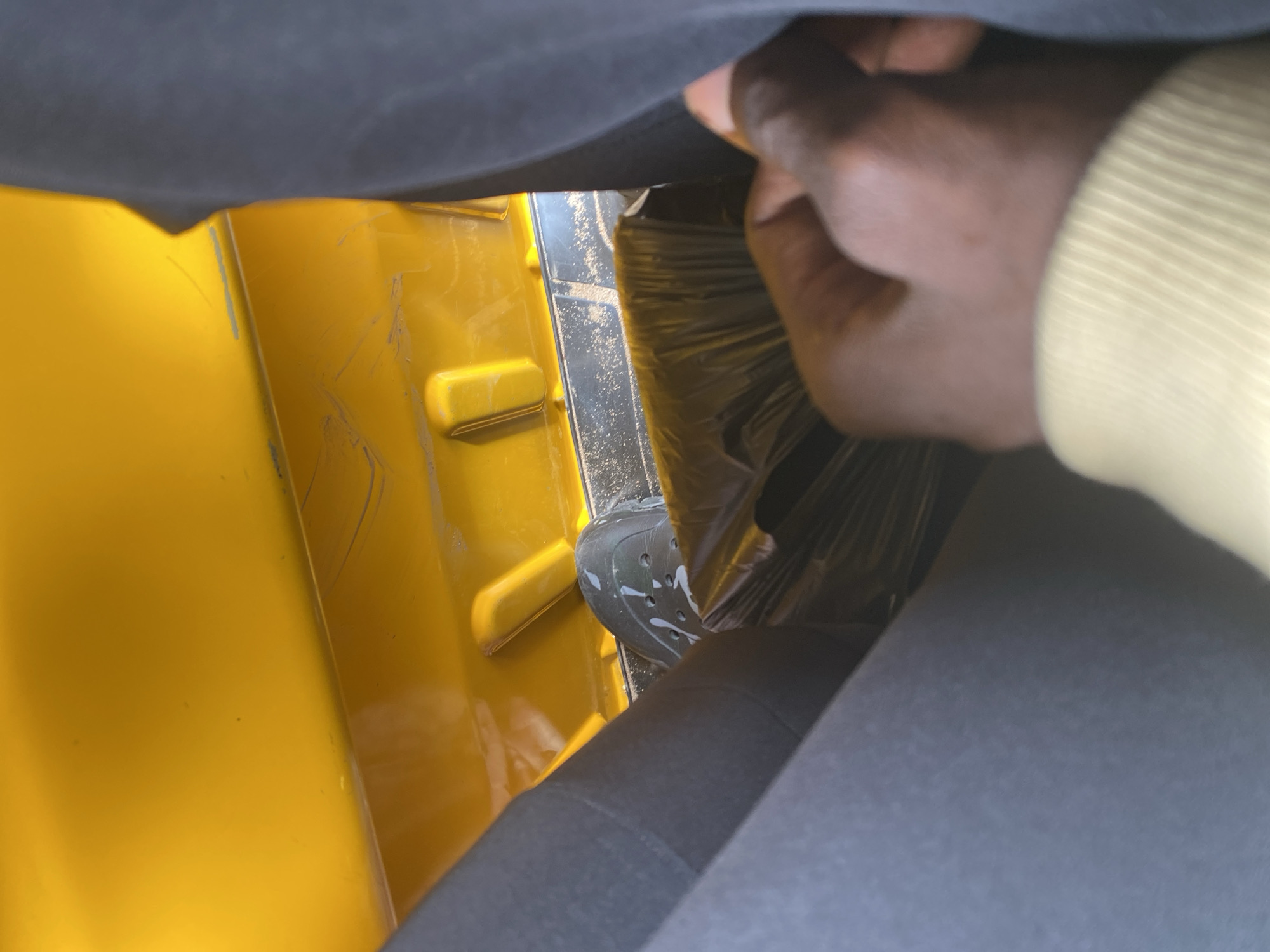 This media file shows a close-up view of a person's hand and foot inside what appears to be a yellow vehicle, possibly a tuk-tuk, in Jalingo, Nigeria. The hand, with dark skin, is reaching towards a black plastic-covered object, possibly a seat or cargo. The person is wearing a light yellow long-sleeved shirt. Their foot is visible, wearing a dark-colored Croc-style shoe with white speckles. The interior of the vehicle is primarily bright yellow with some black accents and what appears to be dust or sand on the surfaces. The overall impression is a candid shot, possibly taken while traveling or loading something into the vehicle. There are no visible text elements, and the lighting suggests it is daytime.