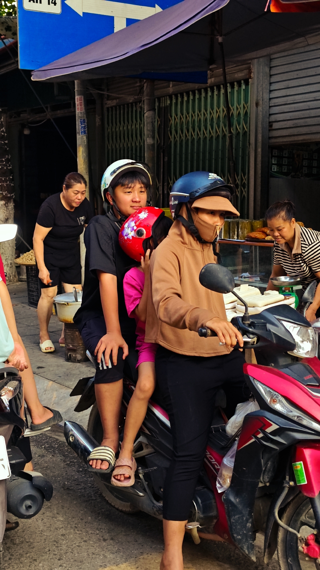 The image depicts a lively street scene in Phố Ràng, Vietnam, likely during a sunny daytime or late afternoon. The foreground is dominated by a red and black Honda scooter carrying three people. A woman, wearing a brown hoodie, black pants, a protective face mask, and a visor under her blue helmet, is in the driver's position with her hands on the handlebars. Behind her sits a child wearing a pink top and a red helmet with a floral design. At the very back is a boy or young man dressed in a black t-shirt, dark shorts with white stripes, and a white helmet. All three have their feet resting on the scooter's footrests, and their helmets indicate typical Vietnamese road safety practices.
In the background, daily activities unfold. To the left, a woman in a black t-shirt and shorts stands near what appears to be a cooking pot or food stall. To the right, another woman in a striped shirt is seen tending to a food display, using a scale to weigh items. The background also features closed green accordion-style metal shop gates, a common sight in urban Vietnamese settings.
Above, a blue road sign with white arrows pointing left and right is visible, partially displaying the text "AH 14." A utility pole also features smaller signs with indecipherable Vietnamese text. The scene captures the everyday bustle of street life, showcasing commuting and small-scale commerce.