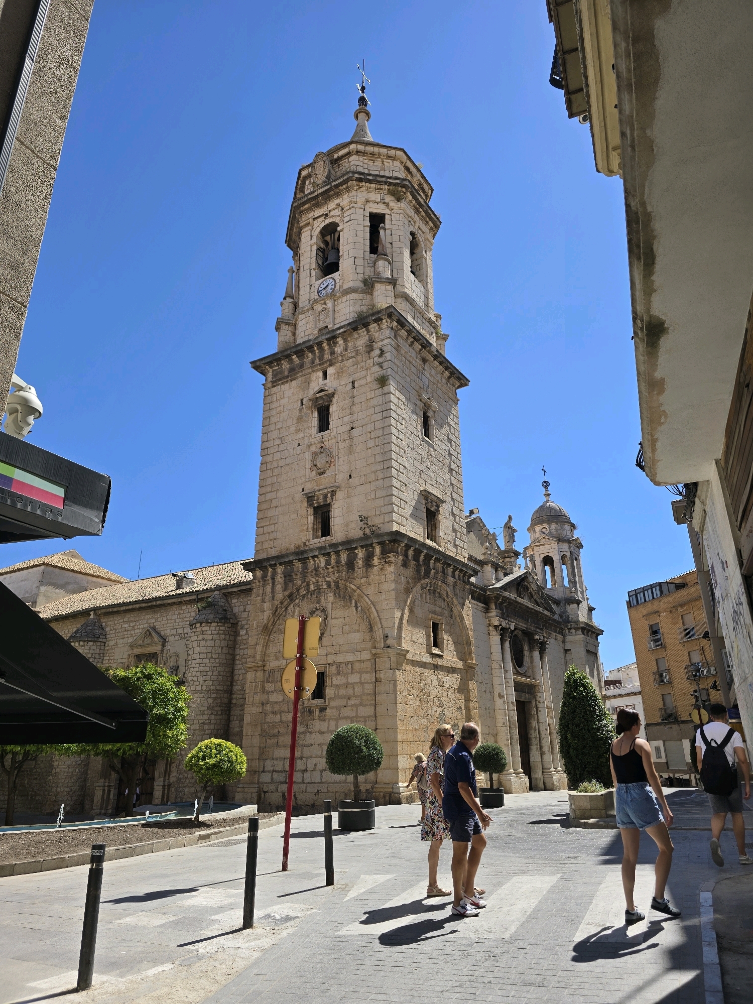The image captures a street scene in Jaén, Spain, dominated by the imposing bell tower and facade of the Jaén Cathedral, a Renaissance architectural masterpiece. The cathedral's stone construction is weathered, hinting at its historical significance. A clear blue sky serves as the backdrop.
In the foreground, a pedestrian crossing is visible, with several individuals traversing it. To the left, a group of three people is walking across the street. The woman on the left wears a patterned dress and sunglasses. Next to her, a man in a dark blue polo shirt and shorts walks with his head turned slightly. A third person, a woman in a floral dress, is slightly behind them and to the left.
To the right of the group, a young woman wearing a black tank top and denim shorts is walking across the pedestrian crossing. She is followed by a man wearing a white t-shirt, dark shorts, and a backpack.
On the right side of the street, a building with balconies and windows is visible. Some of these windows are open, and there are visible air conditioning units. Part of a street sign or informational board is attached to the building.
In the background, to the left of the cathedral, the roof of another building is visible, along with some trees and what appears to be a small fountain area. The street is paved with cobblestones and features bollards to prevent vehicle access in certain areas. The overall atmosphere suggests a sunny day in a bustling urban environment.