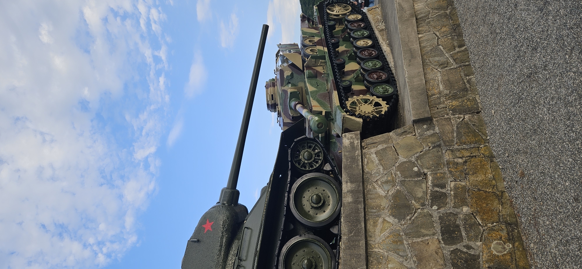 Two historical tanks are displayed on a stone embankment under a partly cloudy blue sky in Svidník, Slovakia. The tank on the left is dark green with a red star emblem on its turret and appears to be a Soviet T-34 model, characterized by its prominent road wheels and distinctive hull shape. The tank on the right is camouflaged with brown, green, and beige patterns and features a German cross emblem on its turret, suggesting it is a German Panzer IV model. Both tanks are positioned with their turrets facing towards the right of the frame. The embankment is constructed of irregular, light-colored stones. In the background, a hill is visible on the right, and the sky is filled with white, scattered clouds against a bright blue backdrop. The ground in front of the embankment is covered with small, dark gray gravel. No people are clearly visible interacting with the tanks in this frame.