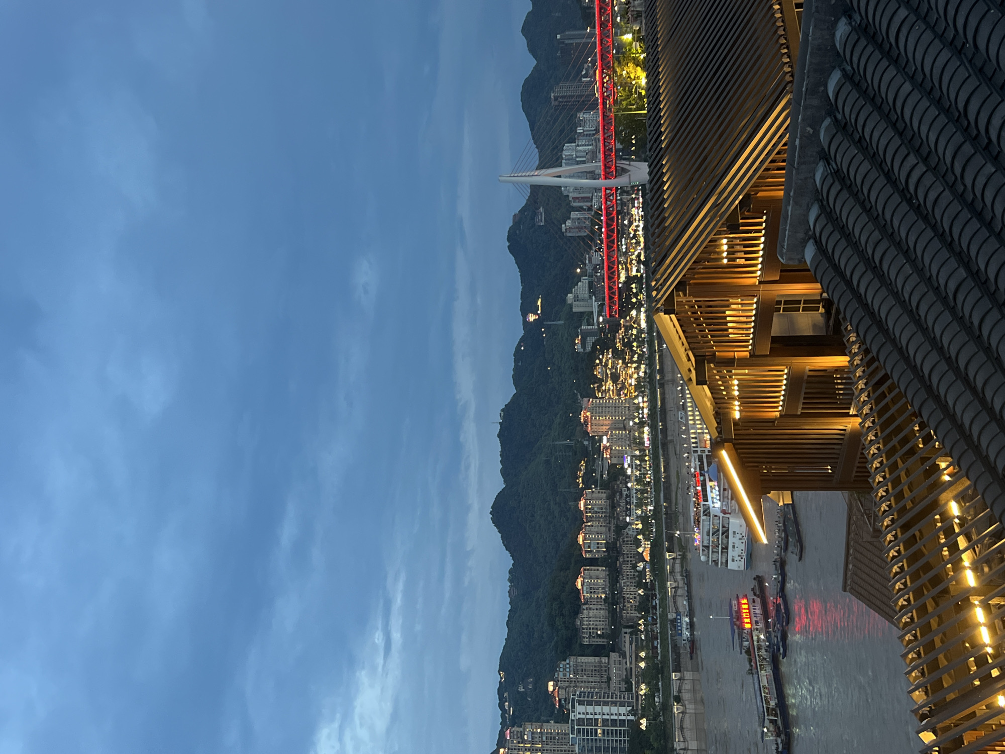 This is an evening photograph taken in the Yuzhong District of Chongqing, China, showcasing a vibrant city skyline along a river. In the foreground, traditional tiled roofs of buildings with wooden structures are illuminated by warm interior lights. The middle ground features the Yangtze River, with several boats and ferries docked or cruising, their lights reflecting on the water. One prominent ferry displays Chinese characters and red lights. Across the river, the city's landscape unfolds with numerous multi-story buildings and residential complexes, many of which are lit up, creating a lively urban glow. Dominating the background are rolling green hills that form a natural backdrop to the urban sprawl. A striking red-lit bridge spans across the river, with a white cable-stayed pylon as its central support. The sky is overcast with deep blue and gray clouds, indicating twilight or a cloudy evening. The overall scene conveys a sense of a bustling, modern city juxtaposed with natural landscape and traditional architectural elements.