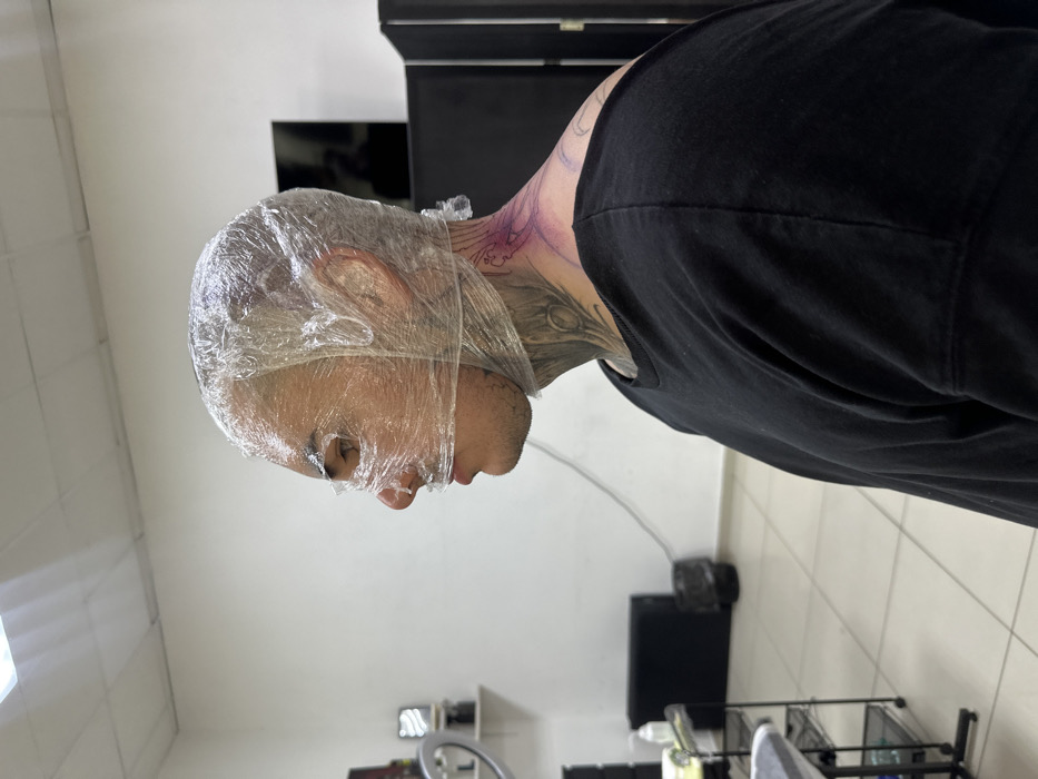 An indoor scene, likely a tattoo studio in Rostov-on-Don, Russia, depicts a male subject in profile, facing left. The subject's head, face, and ears are tightly enveloped in clear plastic cling film, with strategic openings for the eyes and nose. Visible dark-colored tattoos, resembling organic or root-like patterns, cover his neck, extending from beneath his jawline down to his collarbone. A purple stencil outline for a new tattoo is marked on the left side of his upper back and the posterior aspect of his neck. He wears a loose-fitting black t-shirt. The background features plain white walls, a light-tiled floor, and overhead fluorescent lighting. To the left, a multi-tiered black utility cart with various supplies, a ring light on a stand, and a black waste bin are visible. To the right, a dark screen or room divider and a wall-mounted dark rectangular object are present. No other individuals are visible.