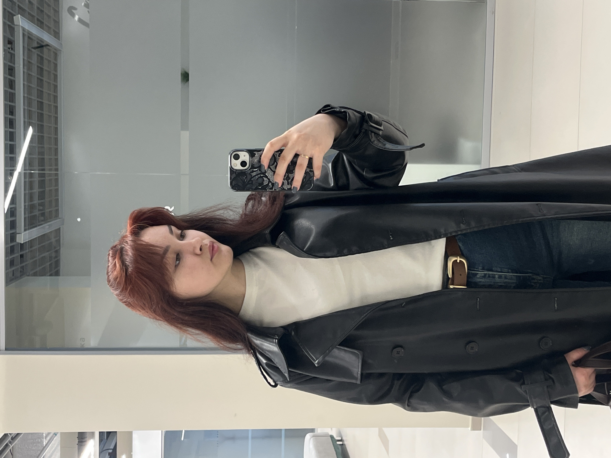 A young woman with reddish-brown hair is taking a selfie in what appears to be a retail environment. She is wearing a white top, blue jeans with a brown belt, and a black leather trench coat. The phone she is holding has a black case with a floral design. The background consists of reflective surfaces, possibly mirrors or glass walls, and a glimpse of the interior architecture suggesting a commercial space. The image lacks specific details to ascertain the exact location in Neftekamsk, Russia.