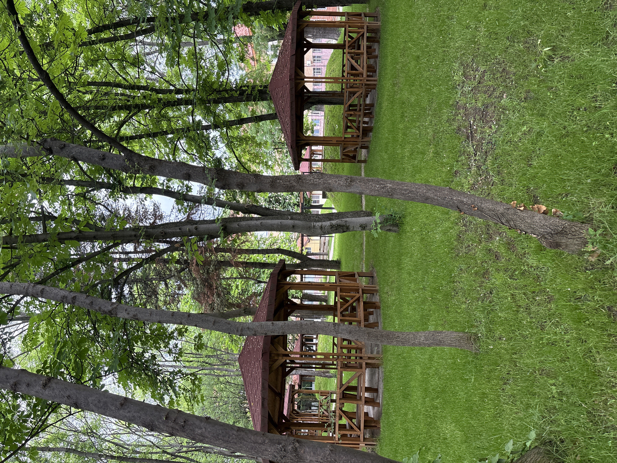 The image depicts an outdoor scene in a park-like setting, potentially in Sofia, Bulgaria. Two wooden gazebos are centrally placed, with a building visible in the background. The ground is covered in grass, and the gazebos are partially obscured by trees. There are no people visible in the image.