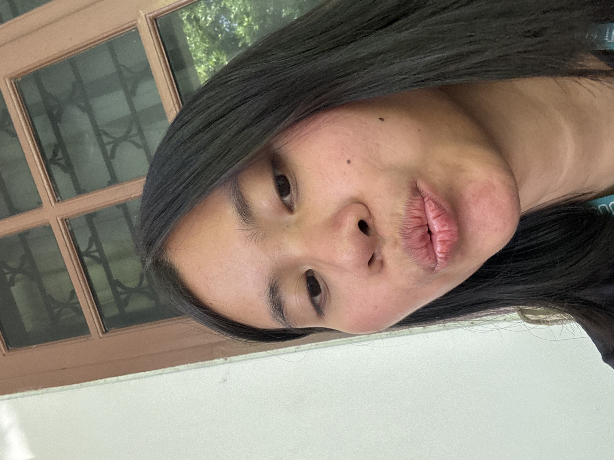 A close-up, selfie-style shot features a young woman with long, dark hair and fair skin puckering her lips. She is positioned in front of a window with brown wooden panes and a dark interior. To her left is a plain, light-colored wall. The overall environment suggests an indoor setting, possibly a home or an office, with a hint of greenery visible through the window, indicating an outdoor space beyond. The subject's gaze is directed towards the camera, and the lighting is natural, highlighting the texture of her skin and lips.