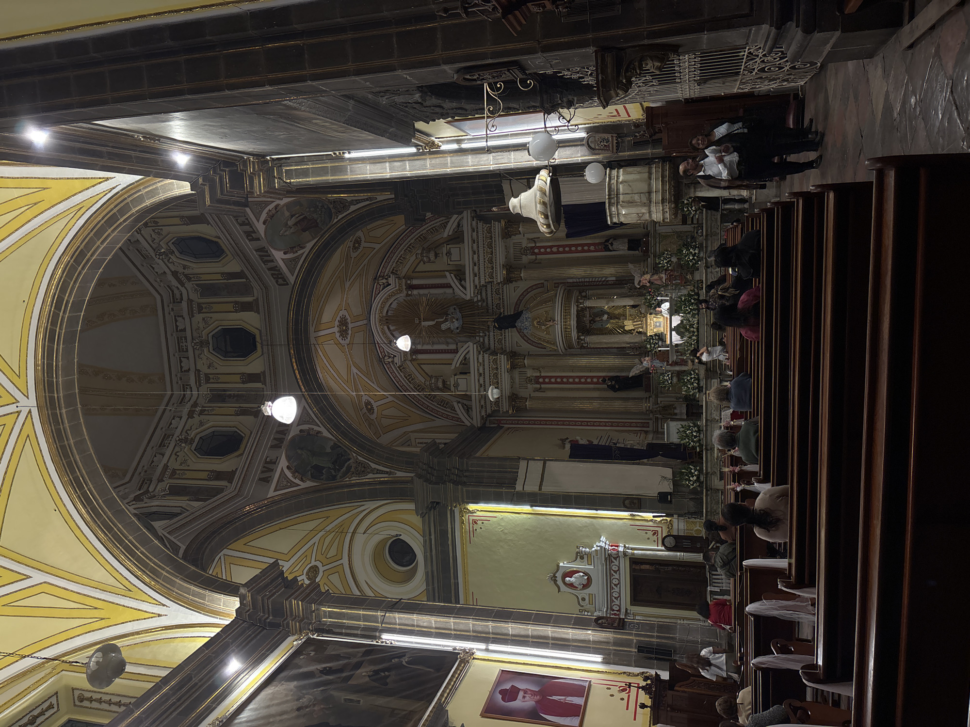 The image shows the interior of a church in Puebla, Mexico. The camera faces the altar. Rows of wooden pews are filled with people, most of whom are sitting. A man and woman stand near the front, facing each other. A large, decorated altar is visible at the far end of the church, adorned with statues and flowers. The church has a high, vaulted ceiling with intricate yellow and gray architectural details, including arches and painted panels. Light fixtures hang from the ceiling. Religious paintings adorn the walls.