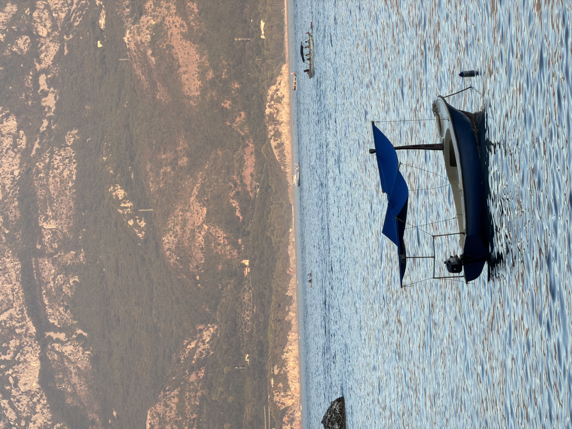 A small, blue motorboat with a blue canopy floats on calm, rippling water. The boat is equipped with an outboard motor. In the background, a densely forested mountain range with rocky outcrops rises behind a coastline dotted with buildings and structures. The sky is a soft, dusky hue, suggesting either sunrise or sunset. Several other boats are visible in the distance, and two people are in a kayak near the shore. The overall scene conveys a serene coastal landscape.
