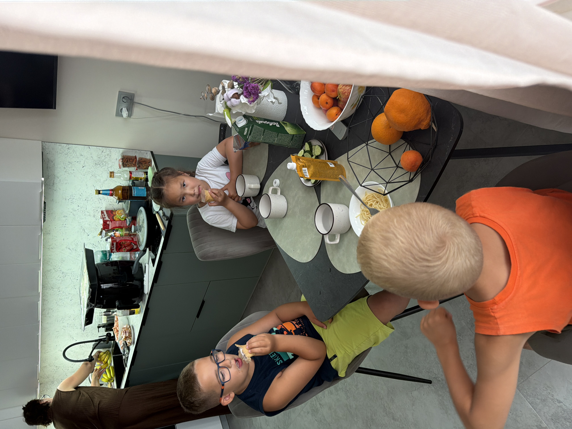 In a domestic kitchen, three children are seated at a dining table. A young boy in an orange t-shirt is facing away from the camera, seemingly engaged in conversation. To his left, a boy wearing glasses and a blue t-shirt is eating from an ice cream cone. Across from him, a girl with braided hair is also eating from an ice cream cone. The table is set with plates of spaghetti, cups, a carton of juice, a bowl of fruit, and sliced cucumbers. In the background, a woman is standing at the kitchen counter, preparing food. The kitchen appears modern and well-equipped, with a sink, stovetop, and an air fryer visible. This scene is set in Novorossiysk, Russia.