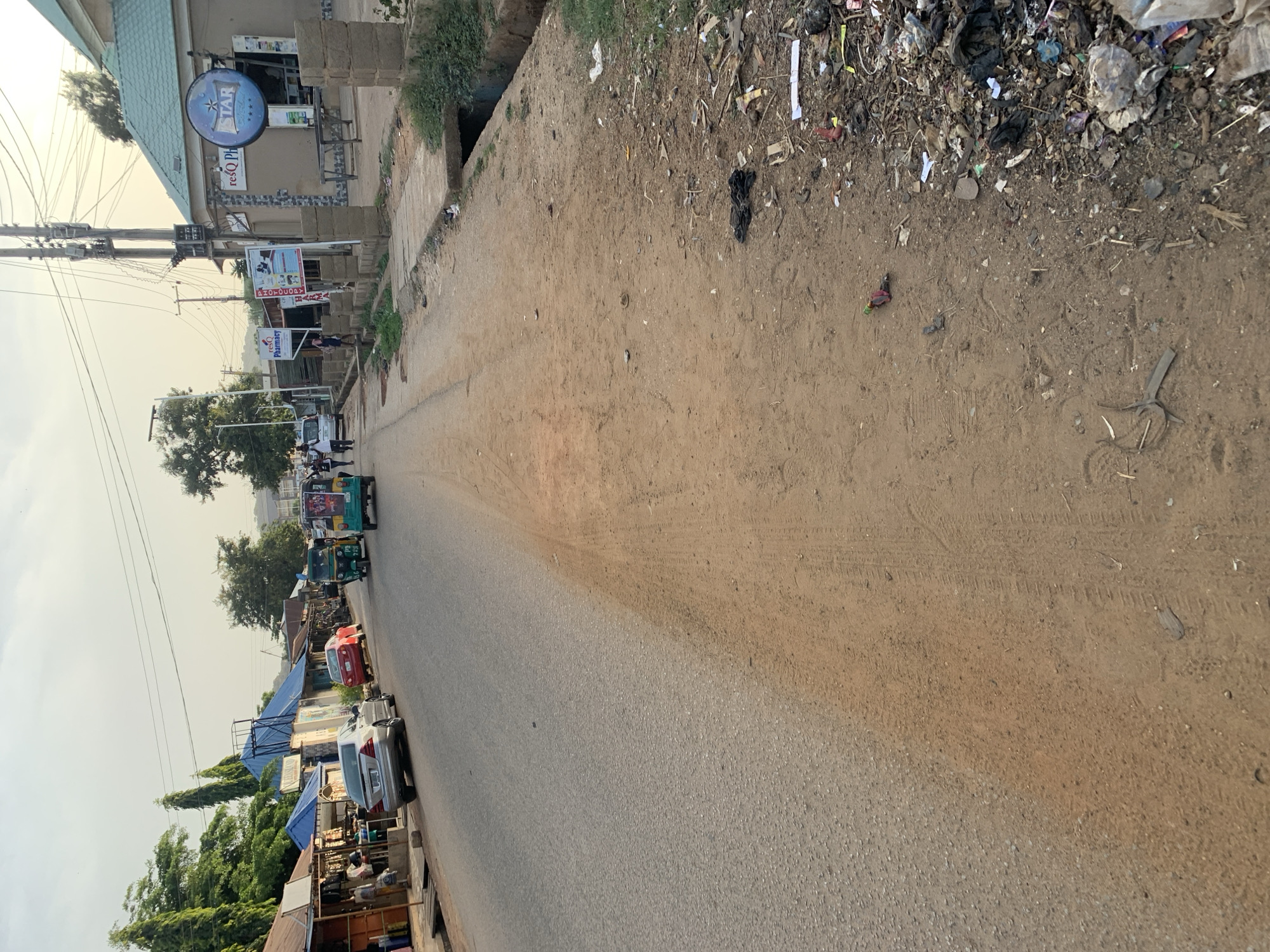 The image shows a street in the New Stadium area of Nigeria. The street runs from the bottom to the top, curving slightly to the left. The left side of the street features buildings, including a "reSQ Pharmacy" and a store with a "STAR" sign. Utility poles with wires run along this side. The right side has a dirt shoulder with trash and debris, including sticks, paper, and plastic waste. Several vehicles, including cars and auto-rickshaws (Tuk-Tuks), are parked or traveling along the left side of the street. People can be seen walking and standing in the distance. The street surface itself appears to be paved. The sky is hazy.