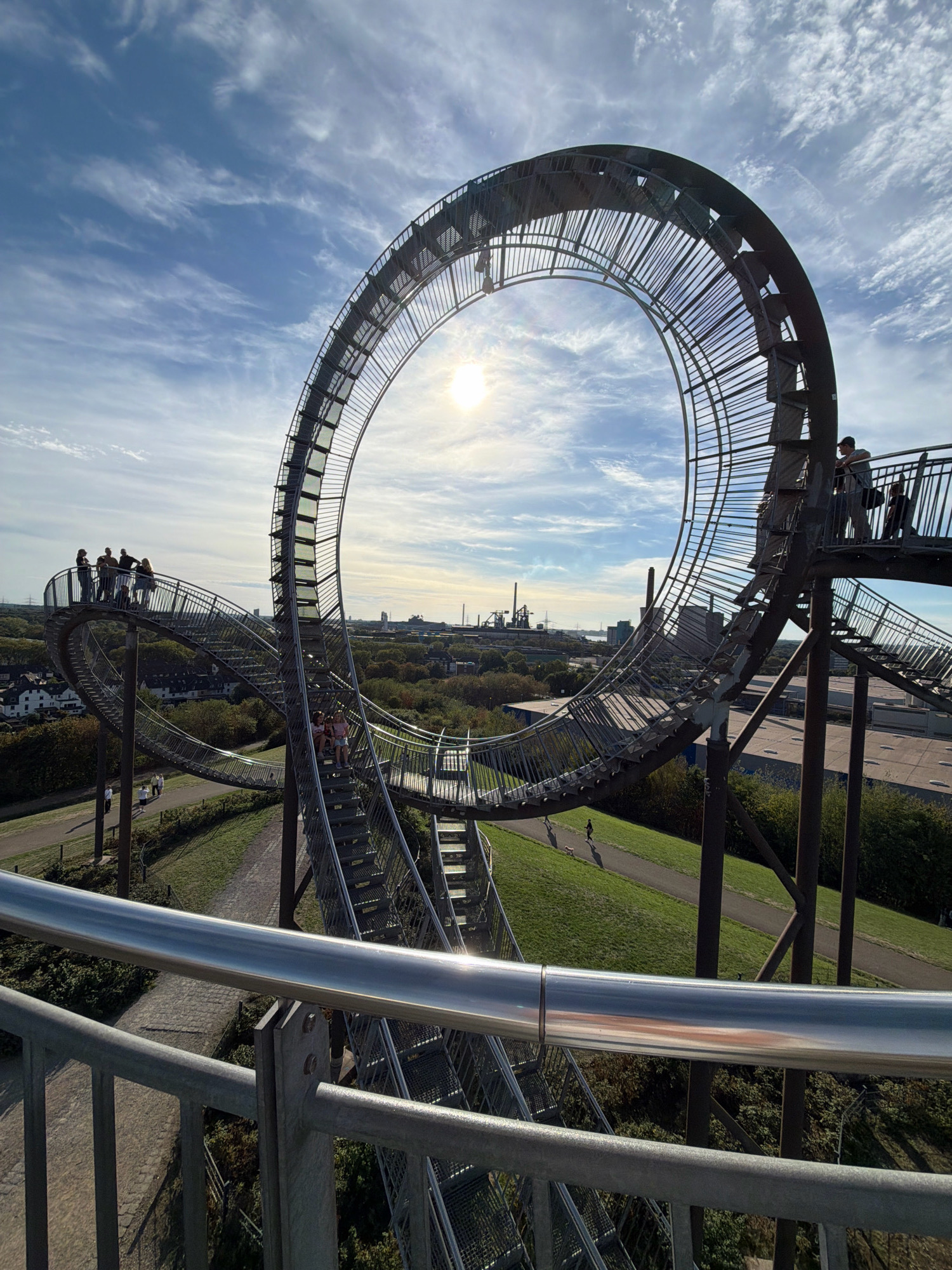 The image captures the "Tiger and Turtle – Magic Mountain" sculpture in Duisburg, Germany, under a partly cloudy sky. The sculpture, a large-scale roller coaster-like structure made of steel, dominates the frame, featuring a prominent loop. Several individuals are visible on and around the sculpture, some standing on platforms and walkways, others ascending or descending the stairs. The surrounding environment includes green, grassy slopes, trees, and in the distance, urban buildings and industrial facilities, characteristic of the industrial landscape of the Ruhr area. The sun is visible in the sky, casting a bright light. The foreground shows a metal railing.