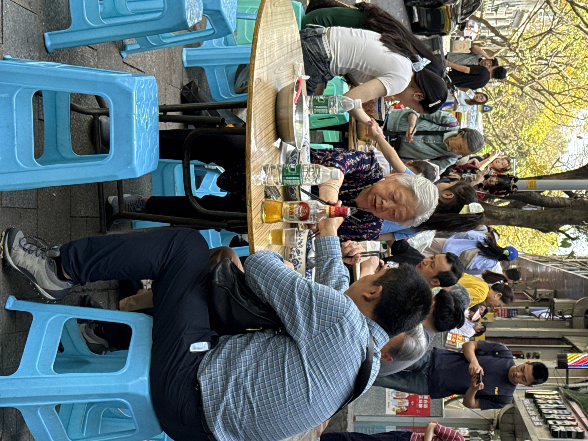 A group of people are seated at a long, oval wooden table, likely eating a meal. The table is covered with bowls, bottles of water, and a bottle of alcohol. They are seated on low, blue plastic stools. The location is in the Yuzhong District, China, as evidenced by Chinese writing and the general ambiance of the street scene. A sidewalk cafe setting, as passersby are walking past. Some people are looking at their phones. Trees overhang the street. There are store front signs in Chinese along the street.