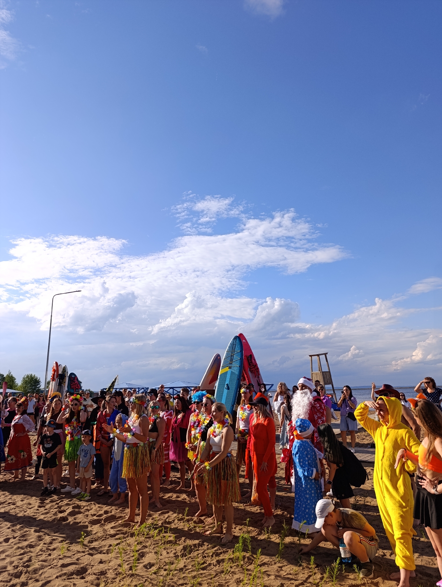 In Chaikovsky, Russia, on a sandy beach under a bright blue sky with scattered clouds, a diverse group of people is gathered near the water. Many individuals are dressed in festive and colorful costumes, including leis, hula skirts, and a Santa Claus outfit. Several surfboards stand upright behind the crowd, suggesting a possible water sports event or festival. The atmosphere appears lively and celebratory, with people of various ages interacting and posing for photos. The background shows a calm body of water and a distant shoreline with trees and some structures. A street lamp stands to the left of the scene.