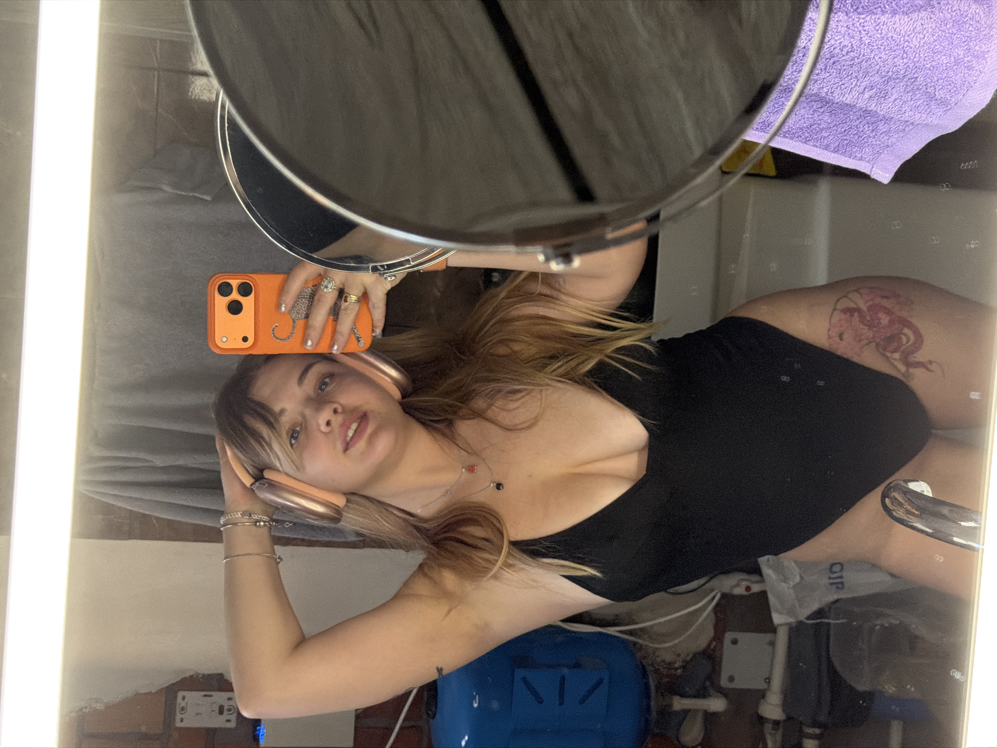 A mirror selfie depicts a young woman, partially clad in a black top or bodysuit, captured in an indoor setting that appears to be a bathroom or dressing room. The image is rotated 90 degrees counter-clockwise. The woman's head is positioned towards the left side of the frame, and her body extends towards the right. She is holding an orange smartphone, featuring a triple-camera setup and what appears to be an orange case, in her right hand to take the photo of her reflection. Her left arm is bent, with her hand resting near the gold-colored over-ear headphones she is wearing.
She has long blonde hair, possibly with bangs or a fringe, and a light complexion, looking directly at the camera with a neutral to slightly alluring expression. Notable details include several rings on her right hand, multiple thin silver bracelets on her left wrist, and a delicate necklace adorned with small red and black beads. A prominent tattoo of a red or pink mythical creature, possibly a dragon or similar design, is visible on her right upper thigh/hip area.
The reflected background reveals a bright fluorescent light fixture running vertically along the left side of the frame. Towards the upper right, a partial view of a larger circular mirror is visible. Further in the background, elements like a folded purple towel, various pipes, cables, and a blue plastic container with some indistinct, reversed text (potentially "OIP") can be seen. The scene conveys a personal and casual moment.