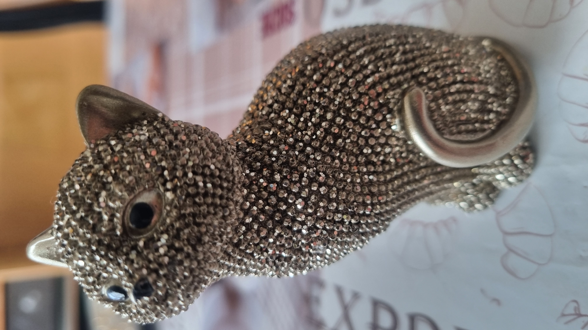 A close-up, eye-level shot focuses on a decorative, silver-colored cat figurine covered in small, sparkling rhinestones. The figurine is seated, facing left, with its tail curled around its body. The cat has small black eyes and pointed ears made of plain silver material. The background is blurred, showing a patterned surface and hints of a wooden object. The scene is devoid of human presence and activity, and there is no contextual information to place it within the city of Smila, Ukraine.