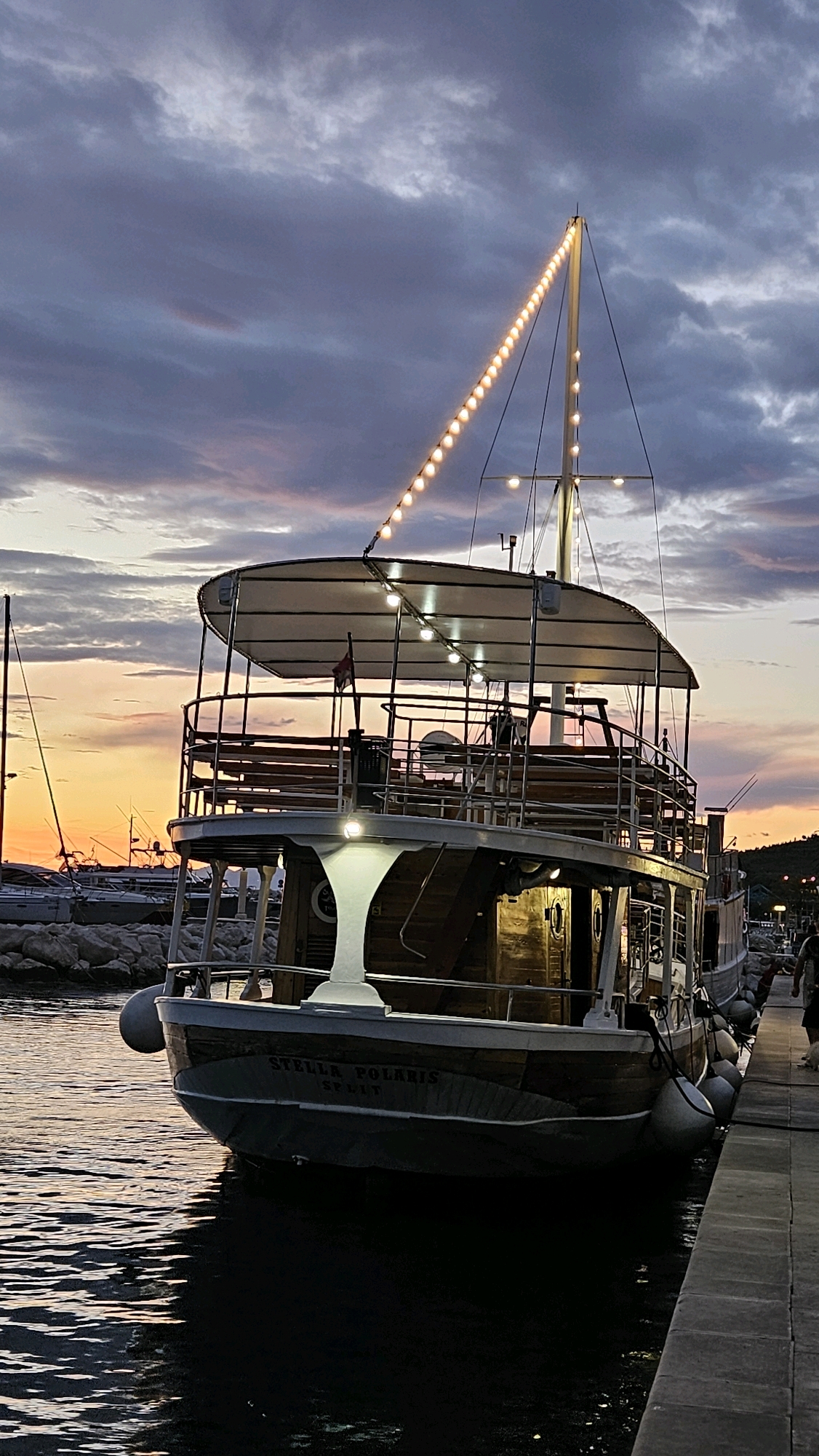 A low-angle, vertical shot captures a docked boat named "STELLA POLARIS SPLIT" at dusk. The boat, with a two-tiered canopy and wooden accents, is illuminated by strings of warm lights along its mast and interior lighting. It's moored at a stone quay, with other boats and a harbor visible in the background. The sky is a blend of deep purples and oranges, indicating sunset or twilight. The water is calm, reflecting the boat's lights and the muted sky. Two white fenders are attached to the side of the boat. The scene appears to be in a harbor, possibly Općina Tučepi, Croatia, given the context.
