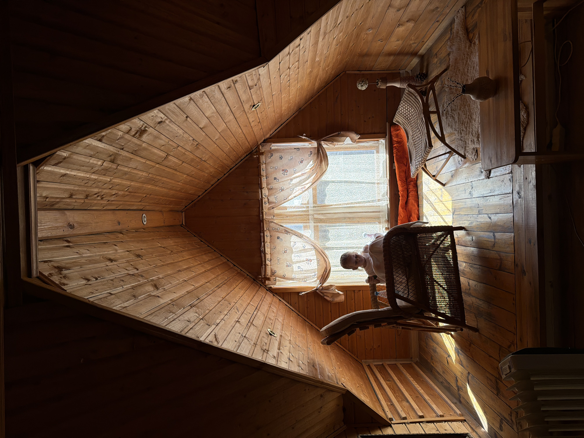 A wood-paneled room, likely an attic, is illuminated by sunlight entering through a window covered with sheer curtains. A young person is visible near the window. A wicker chair with a cushion is present. A table or shelf with decorative objects, including vases and what appears to be a shawl, is on the wall. A humidifier is in the bottom right corner of the frame. The location could be Udelnaya, Russia, given the prompt's instruction.
