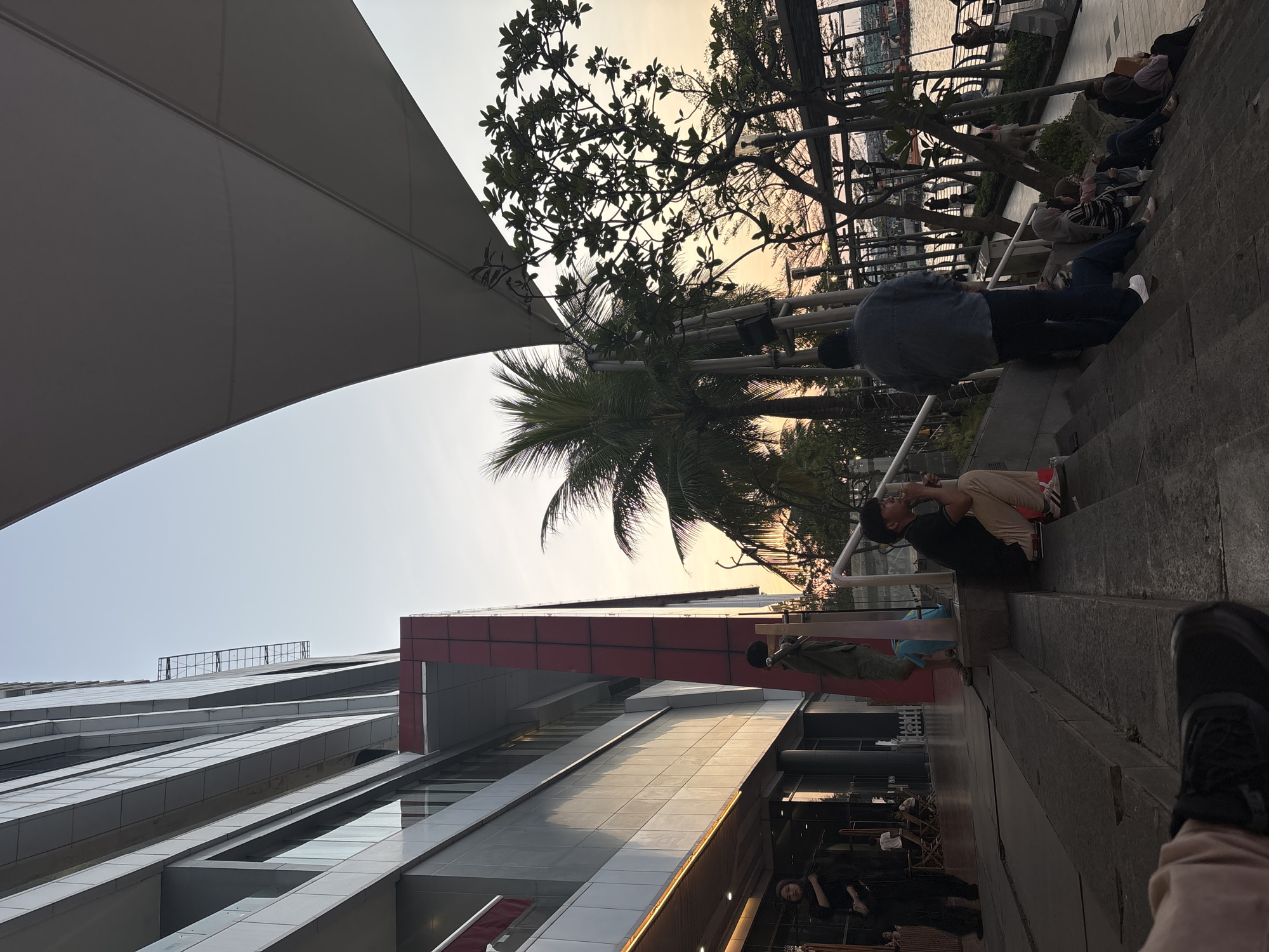 This image captures an outdoor urban scene in North Jakarta, Indonesia, likely within the Pantai Indah Kapuk (PIK) area, during the late afternoon or early evening. The sky transitions from a light blue at the top to orange and yellow tones near the horizon.
To the left, a multi-story modern building with a grey panel facade and large glass windows occupies the upper and mid-sections. A prominent vertical section on the building's right side is clad in reddish-brown material. Sunlight reflects off the lower portion of the building. Below the building, an entrance with an awning is visible. A woman in dark attire and a hijab stands near the entrance, looking towards the camera. Further right on the steps, another individual in a green top stands.
In the mid-ground, a concrete staircase with several steps descends from left to right. A man in a black shirt and tan trousers is crouched on a step, facing right, with his right hand near his face. Below him and to the right, a man wearing a dark denim jacket and dark trousers stands with his back to the camera, looking down the steps. Further down the staircase, a group of approximately five individuals, predominantly women wearing hijabs, are seated on the steps, generally facing right. One person in this group holds a white cup.
The upper right portion of the frame is covered by a large white, angular tensile canopy structure. Several trees are silhouetted against the sunset sky, including a tall palm tree to the left and other leafy trees with visible branches to the right. Beyond the trees and a metal railing, a body of water is visible, with indistinct boats or structures in the far distance on the right. A paved promenade along the water's edge shows several figures of people walking. The bottom left foreground reveals the lower leg and foot of a person wearing light-colored trousers and a black shoe, indicating the camera's low position on the steps.