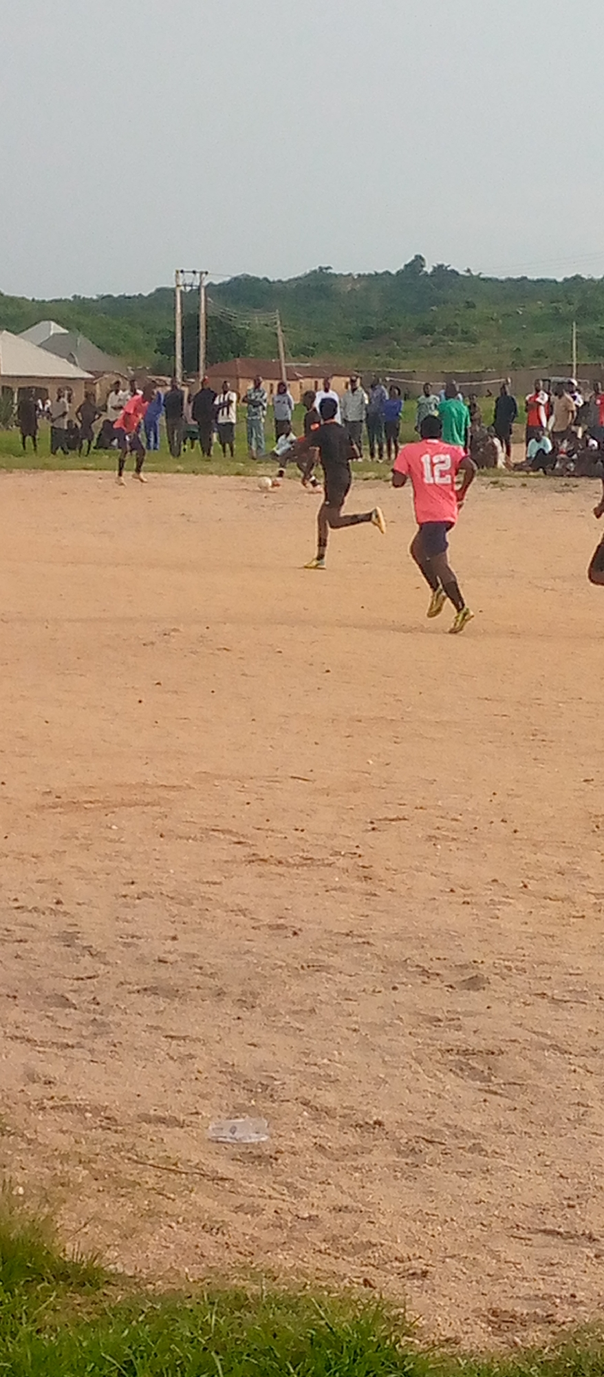 An outdoor, eye-level, medium shot captures a group of people playing soccer on a dusty, uneven field. In the foreground, two young men are actively engaged in the game. One, wearing a pink shirt with the number 12, is running towards the right side of the frame, looking towards a soccer ball. The other, in a black shirt, is also running, his body in motion as if kicking the ball, which is visible near his feet.
In the background, a crowd of spectators is gathered, some standing and others sitting. They are observing the soccer match. The environment appears to be in a rural or semi-urban setting, with some structures and vegetation visible in the distance. The sky is overcast with a hazy light. The ground is covered in dry dirt and sand, with scattered debris. The overall scene depicts a casual soccer game in a community setting.
