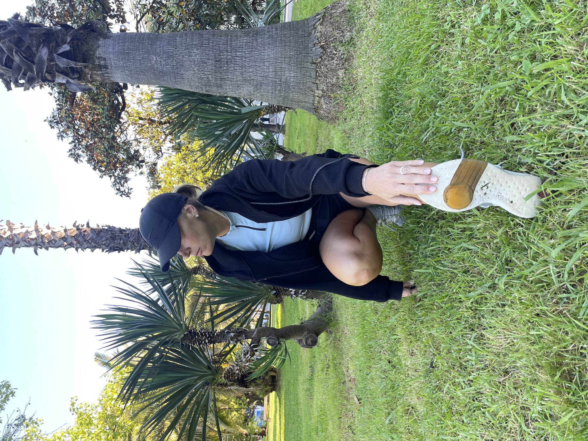 This media file captures a person, likely female, performing a stretch outdoors in a park-like setting. The individual is crouched on green grass, holding their right foot with both hands to stretch their calf or Achilles tendon. They are dressed in athletic wear, including a dark baseball cap, a light-colored long-sleeved top, a dark zippered jacket, and dark shorts. A silver bracelet is visible on their left wrist, along with a ring on their left ring finger. Their light hair is pulled back, partially visible from under the cap.
The scene is brightly lit by natural sunlight, suggesting it is daytime with clear weather. The ground is covered in vibrant green grass. Numerous trees are scattered throughout the background; prominent palm trees with textured trunks and fronds are visible, alongside other leafy trees. The sky is clear and light-colored. On the sole of the right shoe, the brand "Reebok ∆" is discernible. The environment, with its lush greenery and palm trees under a sunny sky, aligns with the characteristics of Sochi, Russia. The image conveys a moment of exercise or warm-up in a pleasant outdoor space.