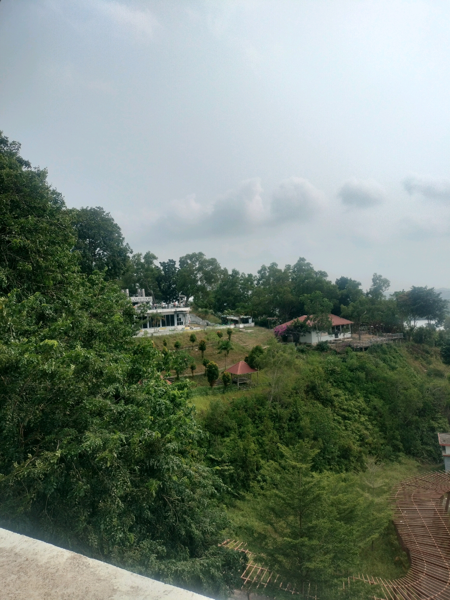 The image depicts a scenic view of a lush, green hillside overlooking a body of water, likely a river or bay, on a cloudy day. Several buildings are situated on the slope, including a white building with a prominent sign that appears to read "GRAND BRIDGE." Another building with a red-tiled roof is visible further down the hill, with an open-air structure nearby. The landscape is characterized by dense trees, well-maintained grassy areas, and winding pathways. In the foreground, a light-colored concrete structure suggests a vantage point or balcony. The presence of a building with a sign and its location on a hillside with amenities suggests it might be a resort, park, or a recreational facility. Based on the architectural style and vegetation, the scene could potentially be located in a coastal or hilly region of Batam, Indonesia, a popular tourist destination known for its natural beauty and resorts.