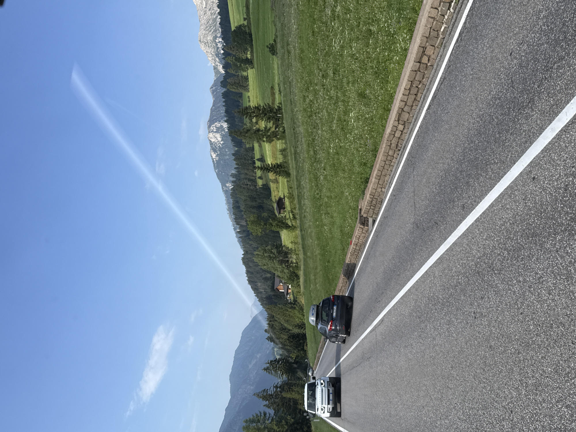 The image depicts a scenic view from a road in San Giovanni di Fassa - Sèn Jan, Italy, on a clear day with a blue sky and scattered clouds. The foreground shows a paved road with white lane markings. Two cars are visible on the road: a white SUV on the left and a dark-colored hatchback with a roof-top carrier on the right. In the middle ground, a lush green meadow slopes upwards, dotted with wildflowers and trees. Further back, a dense forest covers the foothills of majestic mountains, with some scattered houses visible amongst the trees. The mountains themselves are rocky and imposing, with snow-capped peaks in the distance. A faint contrail is visible in the sky, stretching diagonally across the frame. The overall impression is one of natural beauty and tranquility.