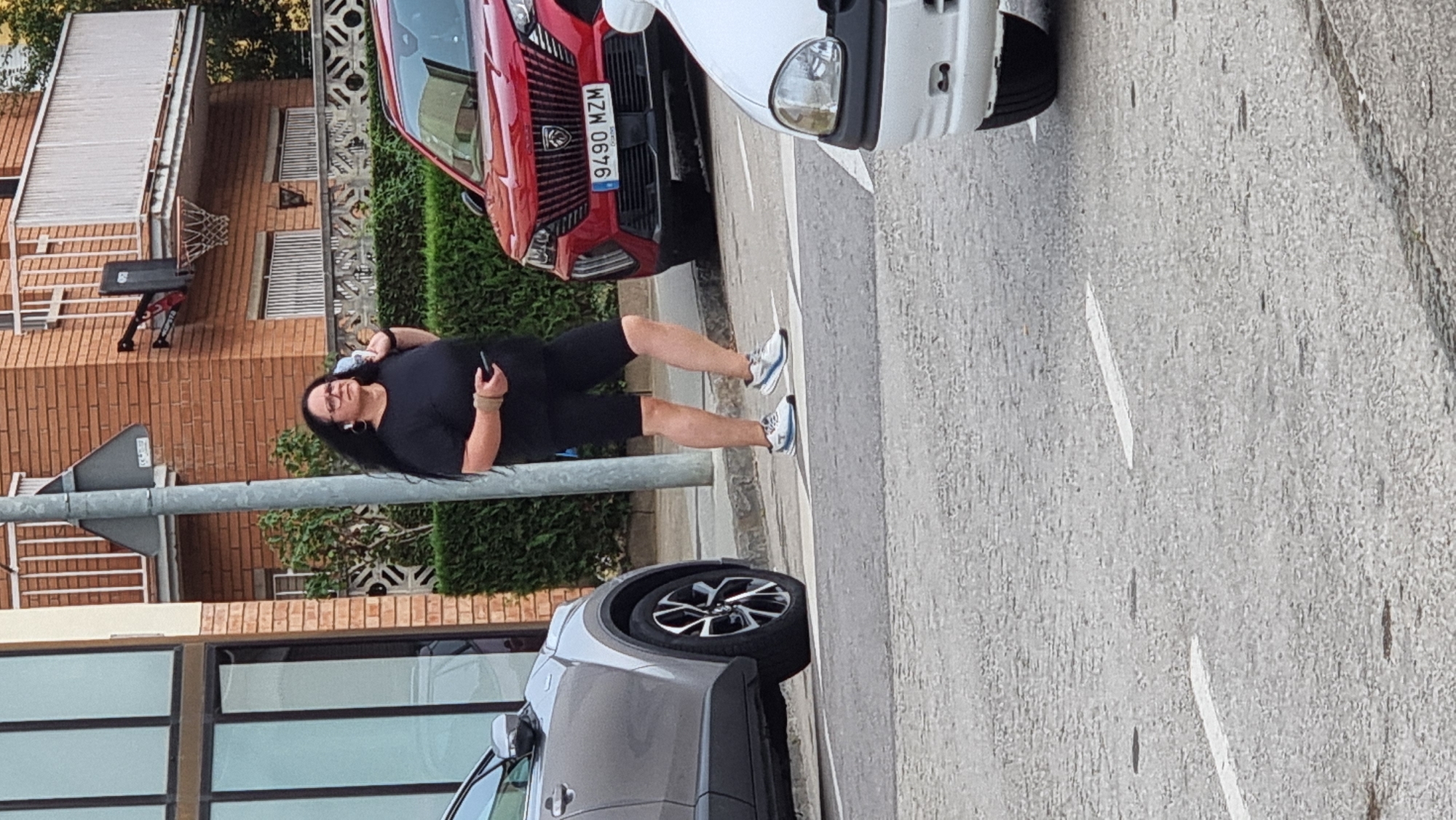 A woman with long dark hair and glasses is standing on a street in Sant Joan Les Fonts, Spain. She is wearing a black t-shirt, black shorts, and athletic shoes. She is holding a mobile phone in her right hand and has a light-colored object slung over her right shoulder. She is looking to her left, with a neutral expression. The woman is standing on a pedestrian crossing, with a grey car to her left and a red SUV to her right. The red SUV has a license plate that reads "9490 MZM". In the background, there is a brick building with a balcony and a basketball hoop attached to the side. There is also a hedge and a street lamp. The weather appears to be clear and sunny.